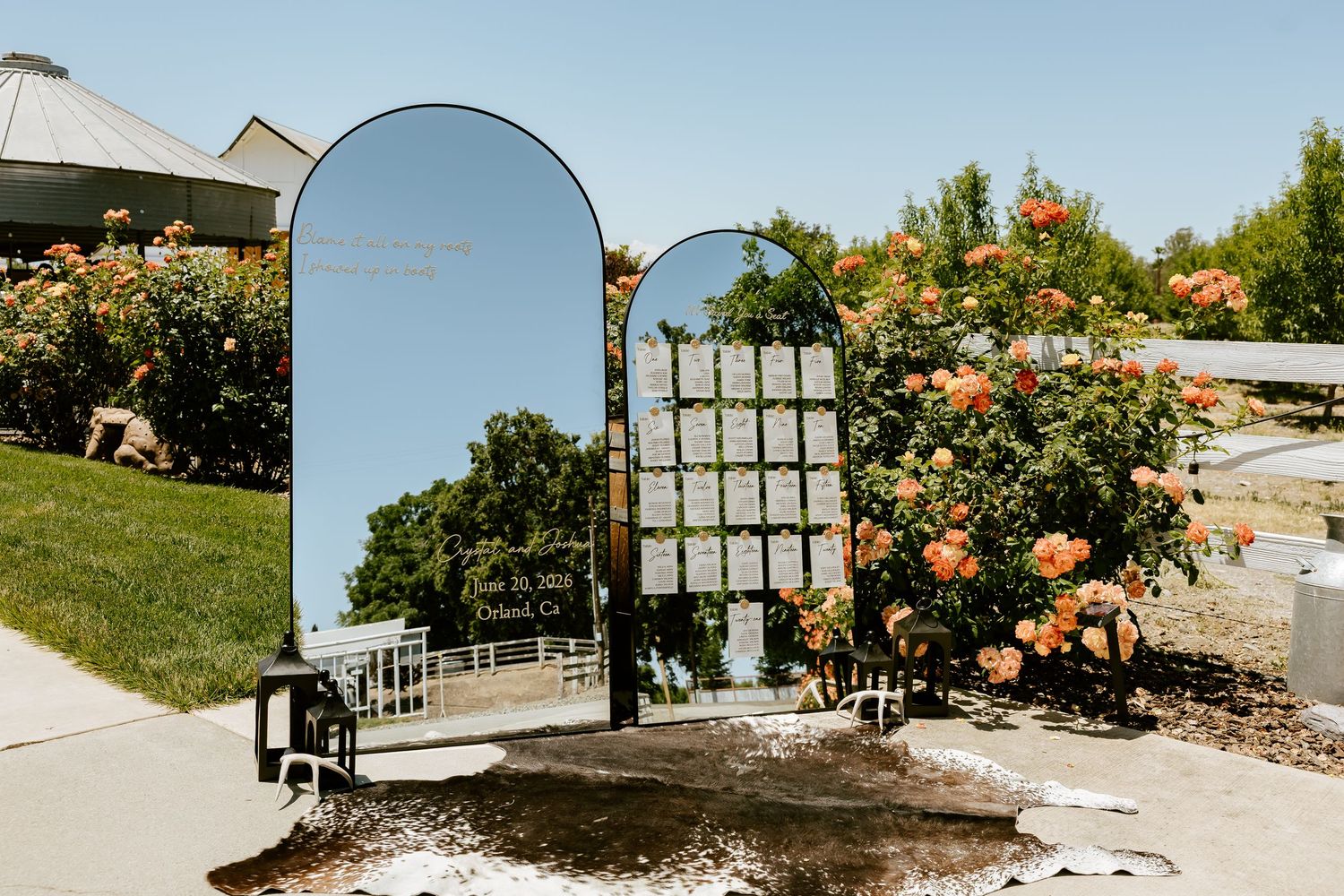 Elegant arched mirrors displaying wedding seating charts outdoors, surrounded by blooming orange roses on a sunny day.