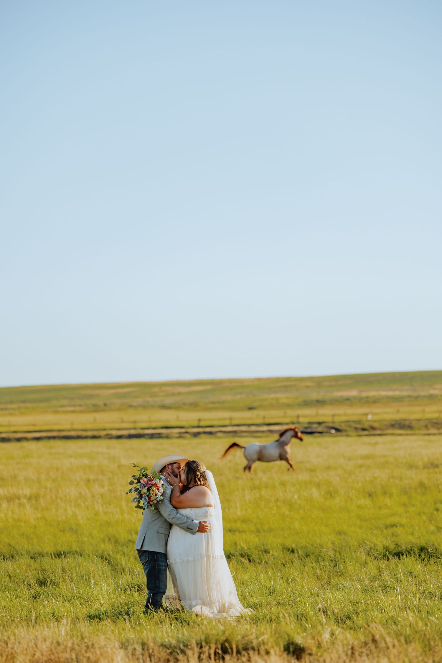 A bride and groom embrace in a green field with a white horse galloping in the background on their wedding day.