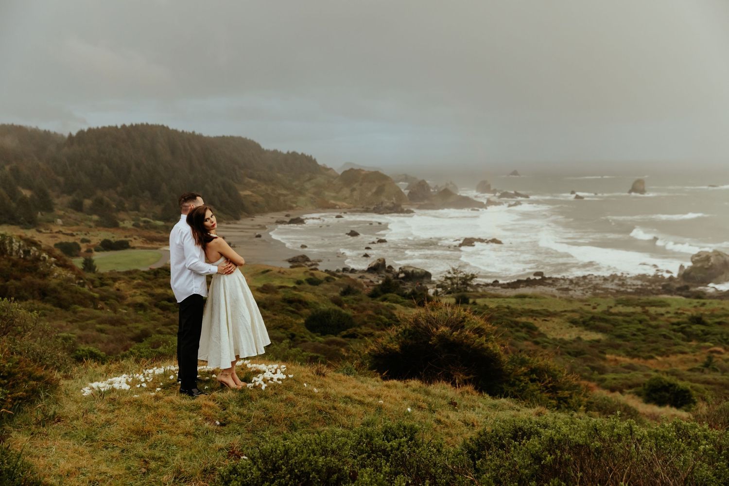A romantic coastal wedding photo showcasing a couple embracing on a cliff overlooking a dramatic rocky shoreline.