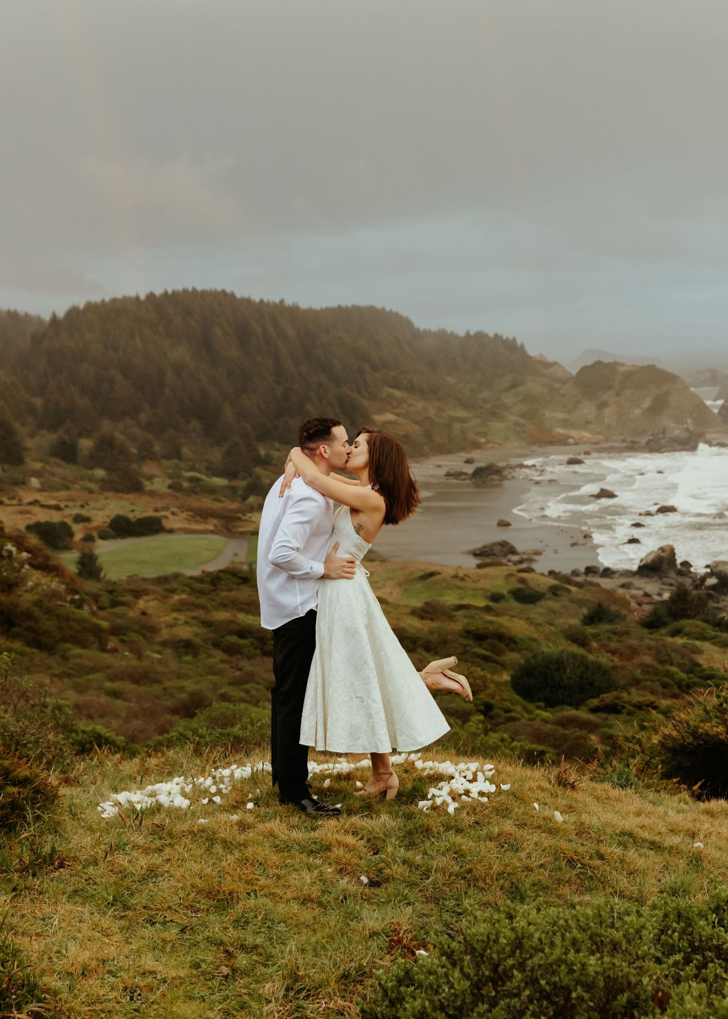 A romantic proposal moment on a scenic coastal cliff overlooking the rocky Pacific shoreline at sunset.