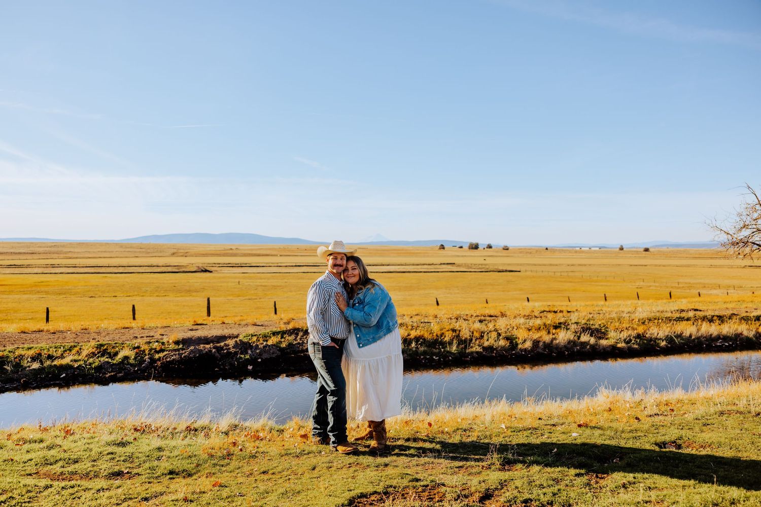 A couple embraces by riverside with golden fields stretching to distant mountains.