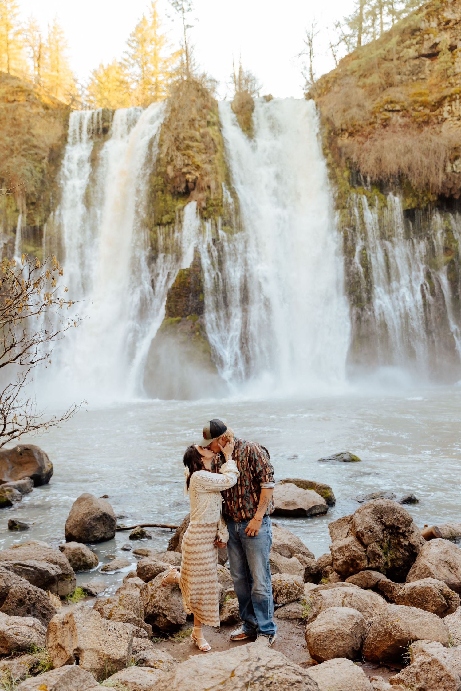 Two people share a romantic moment in front of a majestic cascading waterfall surrounded by autumn trees and rocky terrain.