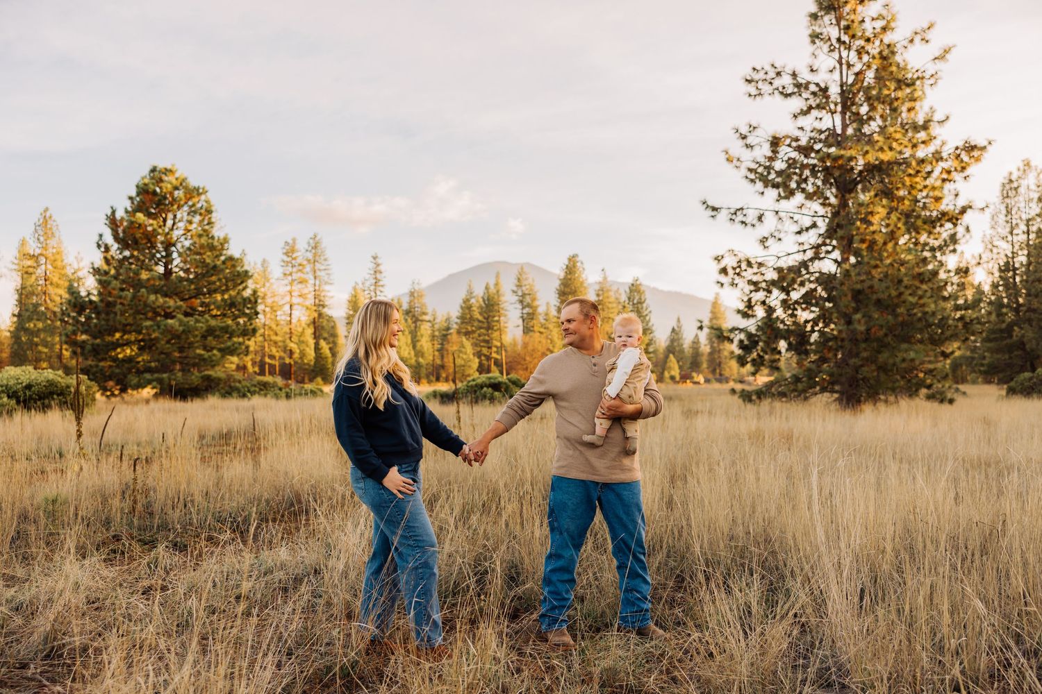 A couple shares a playful moment in a golden autumn meadow with mountains and evergreens in the background.