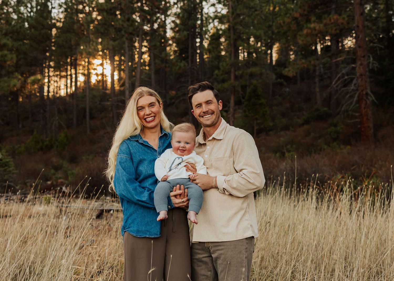 Family poses together in a golden field at sunset with pine trees in the background.