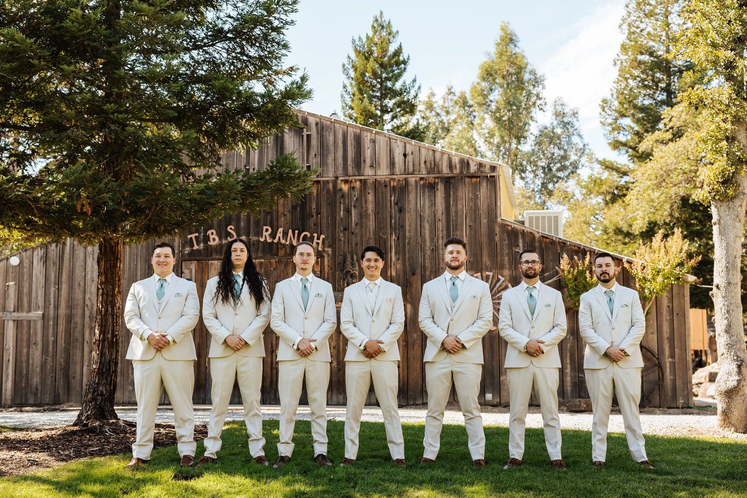 Wedding party in white suits posing in front of a rustic barn at TBS Ranch outdoor venue.