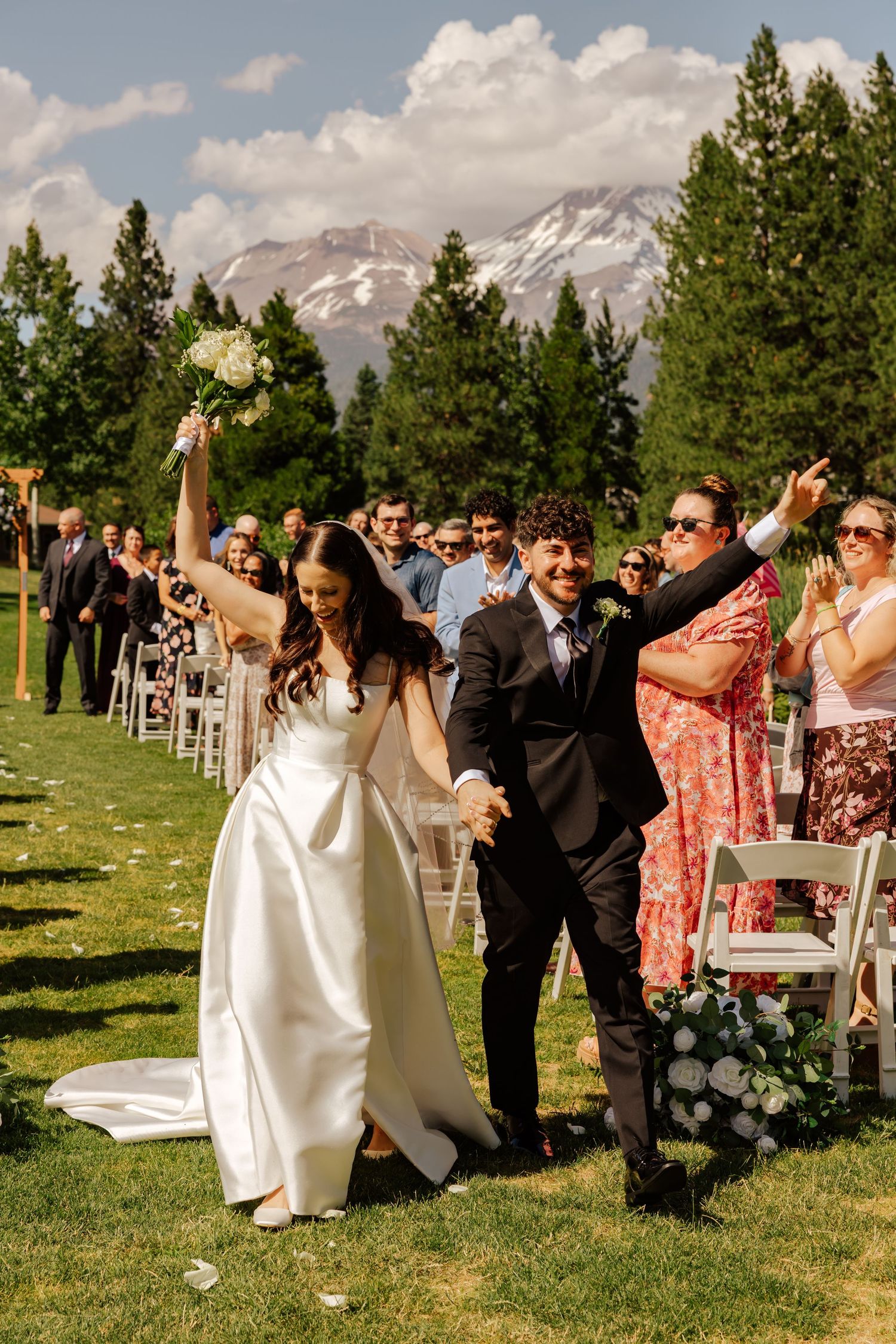 Bride and groom walk joyfully down outdoor wedding aisle with mountain backdrop and cheering guests.