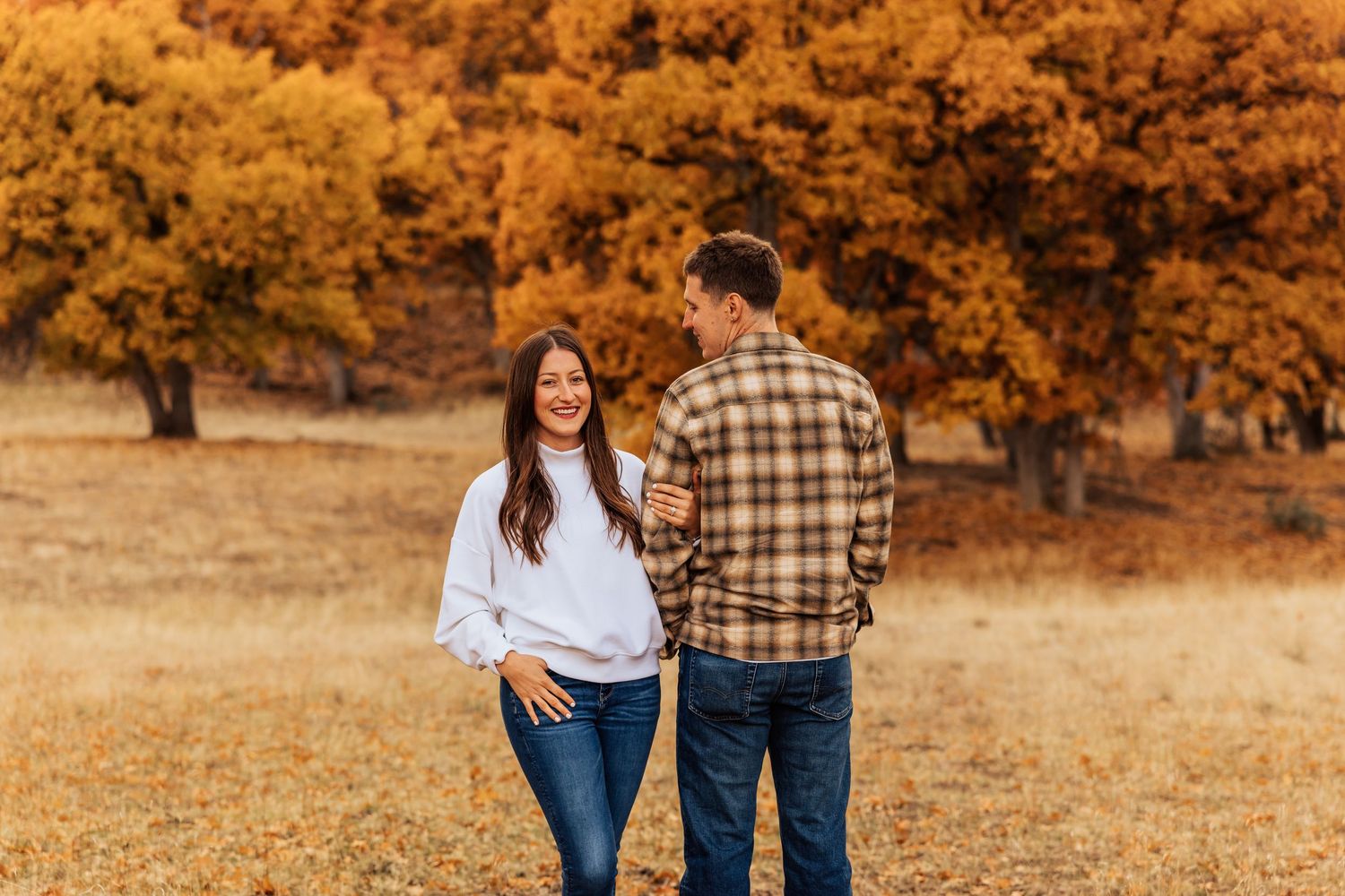 Couple walking through an autumn landscape with golden trees and dry grass.