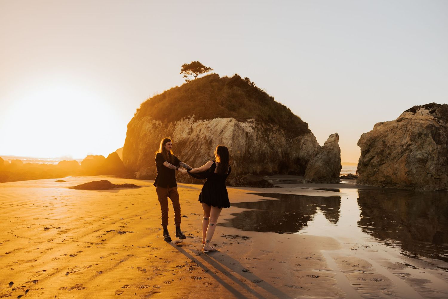 Two silhouettes walking on sandy beach at sunset with large rock formations and ocean waves.