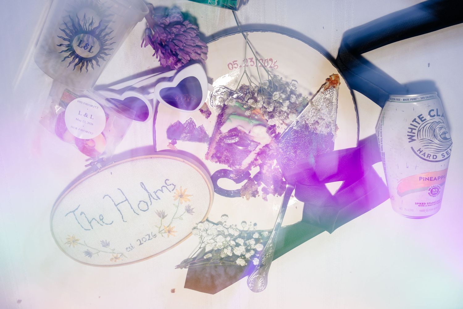 Dreamy overexposed flat lay featuring The Holms sign, dried flowers, and White Claw can in soft purple hues.