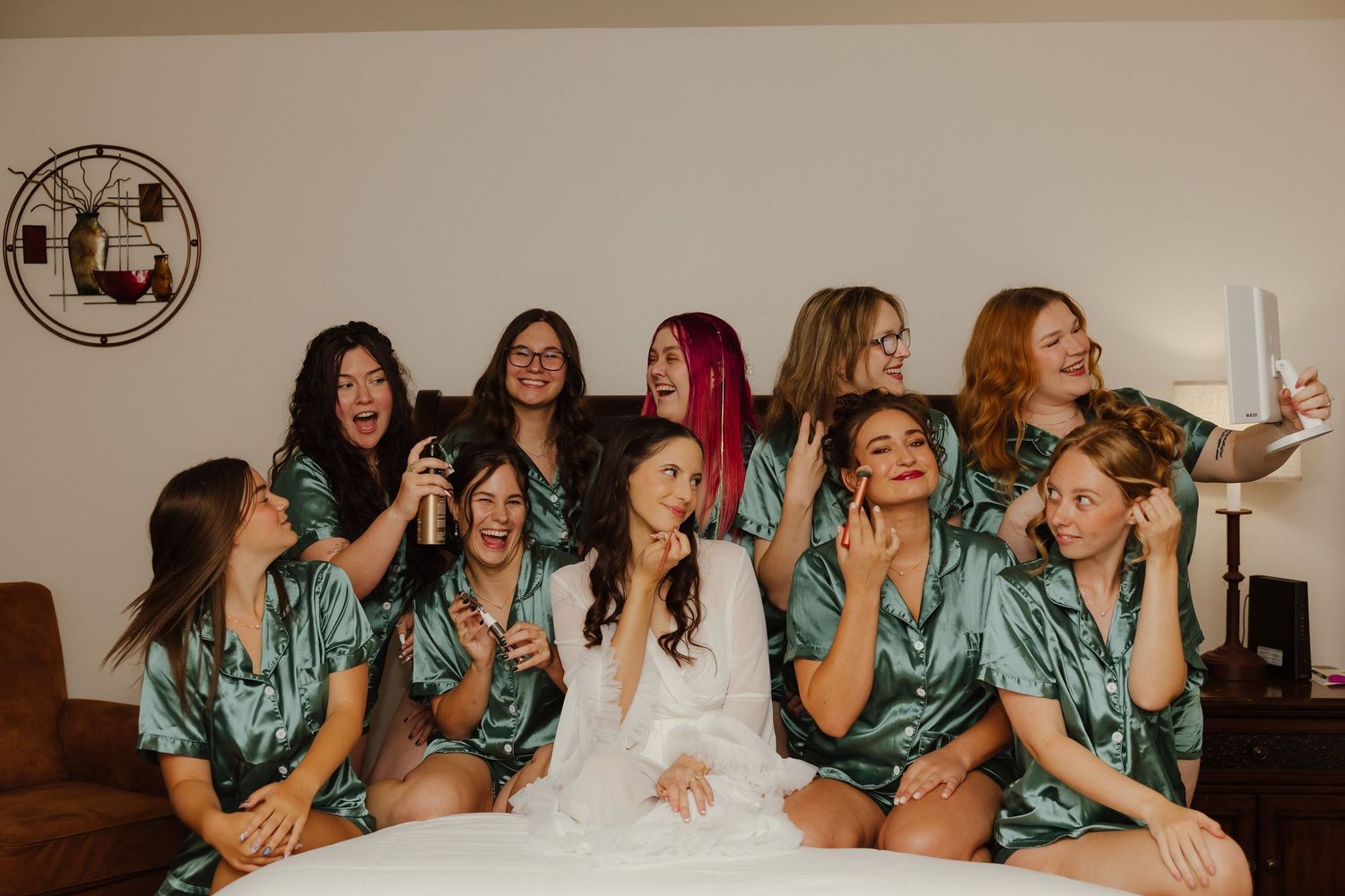Bride in white robe takes selfie with bridesmaids in matching green satin robes on bed during wedding morning prep.