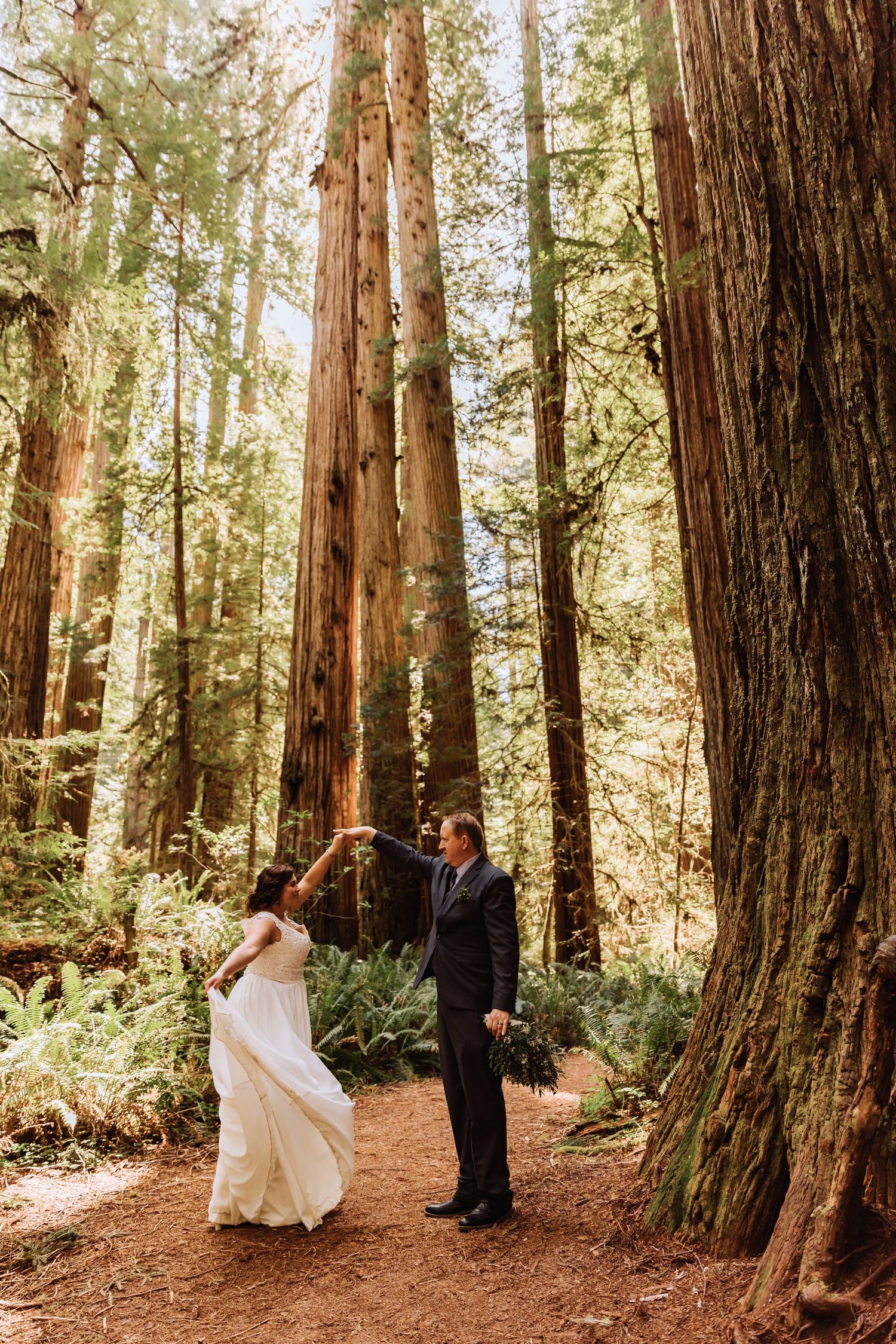 Wedding couple poses among massive redwood trees on a rustic forest trail.