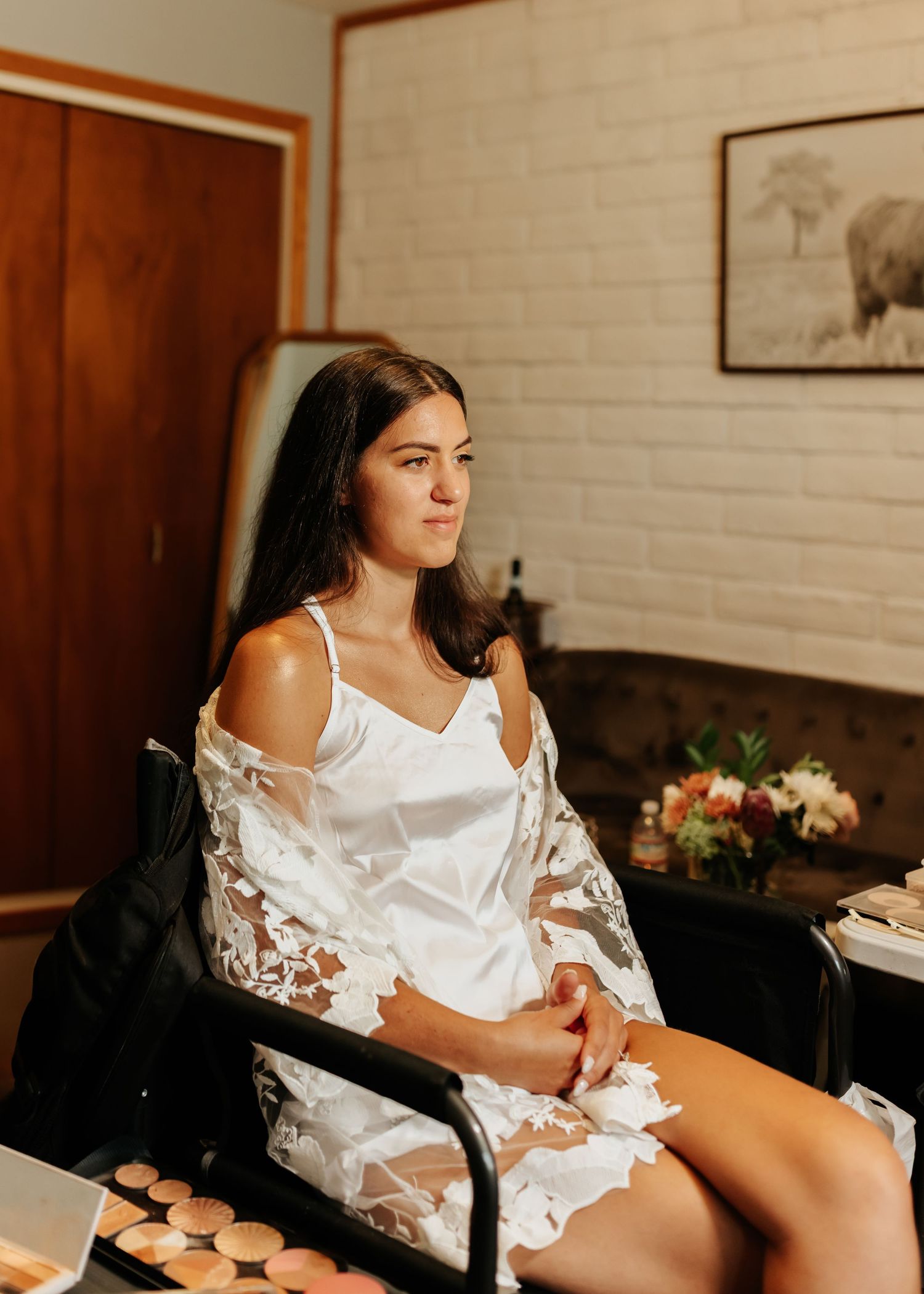 Bride getting ready in a white robe with lace details while sitting in an elegant indoor setting.