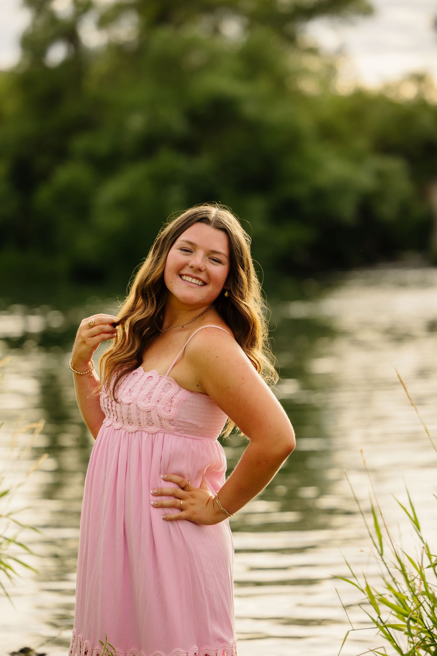 Young woman in pink sundress smiling by a serene lake with lush green trees in the background.