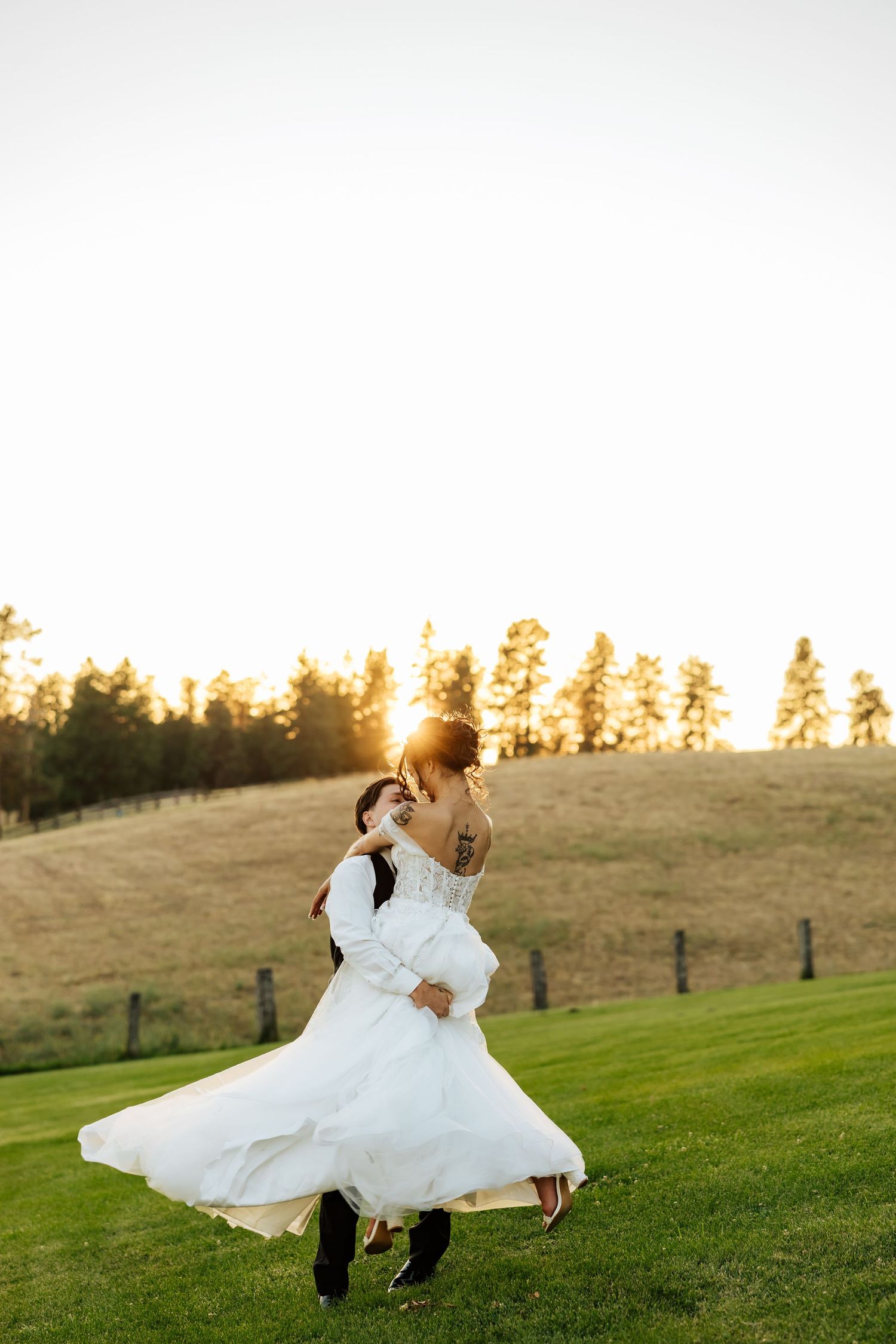 Groom lifts bride in white gown at golden sunset on green hillside, trees in background.