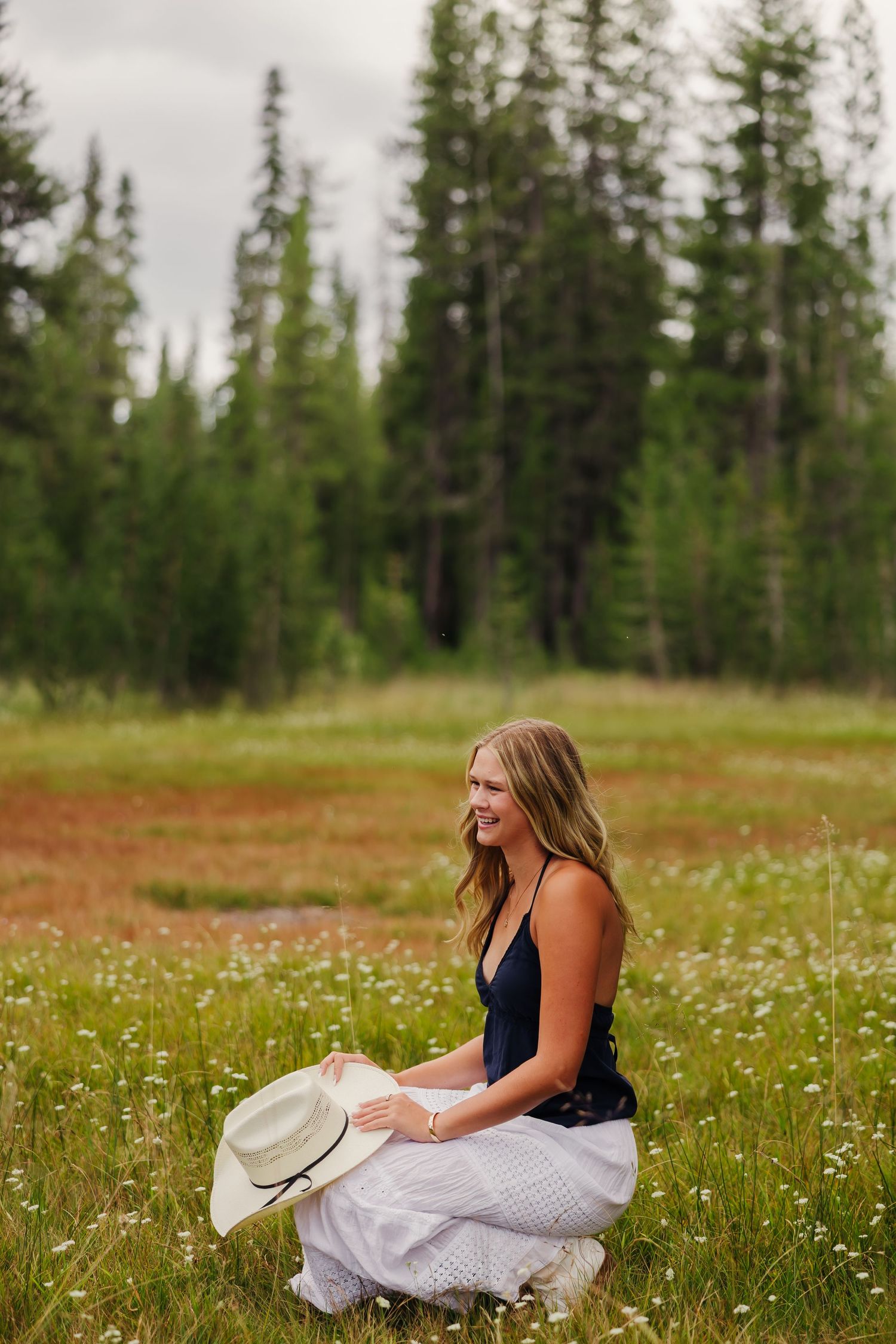 Young woman in white skirt holding a straw hat, sitting in a wildflower meadow with pine trees behind her.