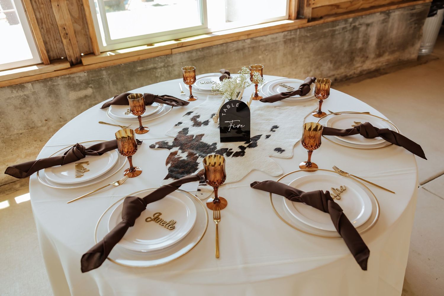 Elegant round wedding reception table with gold goblets, dark napkins, white plates, and a cowhide centerpiece.