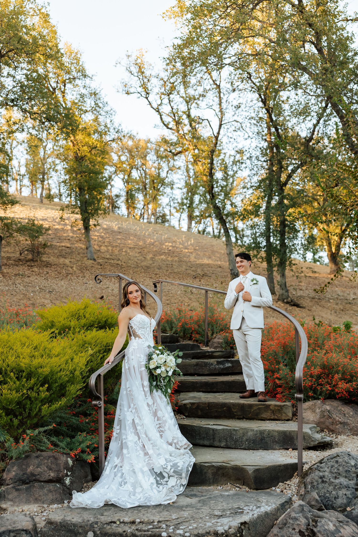 Bride in lace gown holding bouquet stands on stone steps with groom in white suit behind her, surrounded by autumn trees.