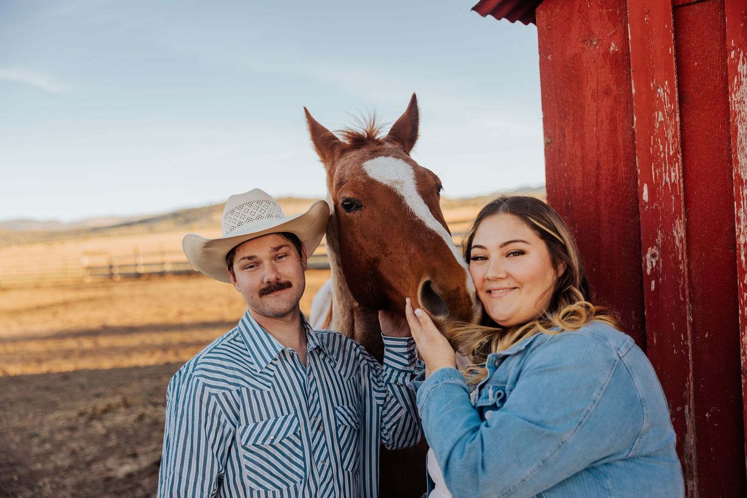 A romantic moment captured near a red barn with horses in the background at sunset.
