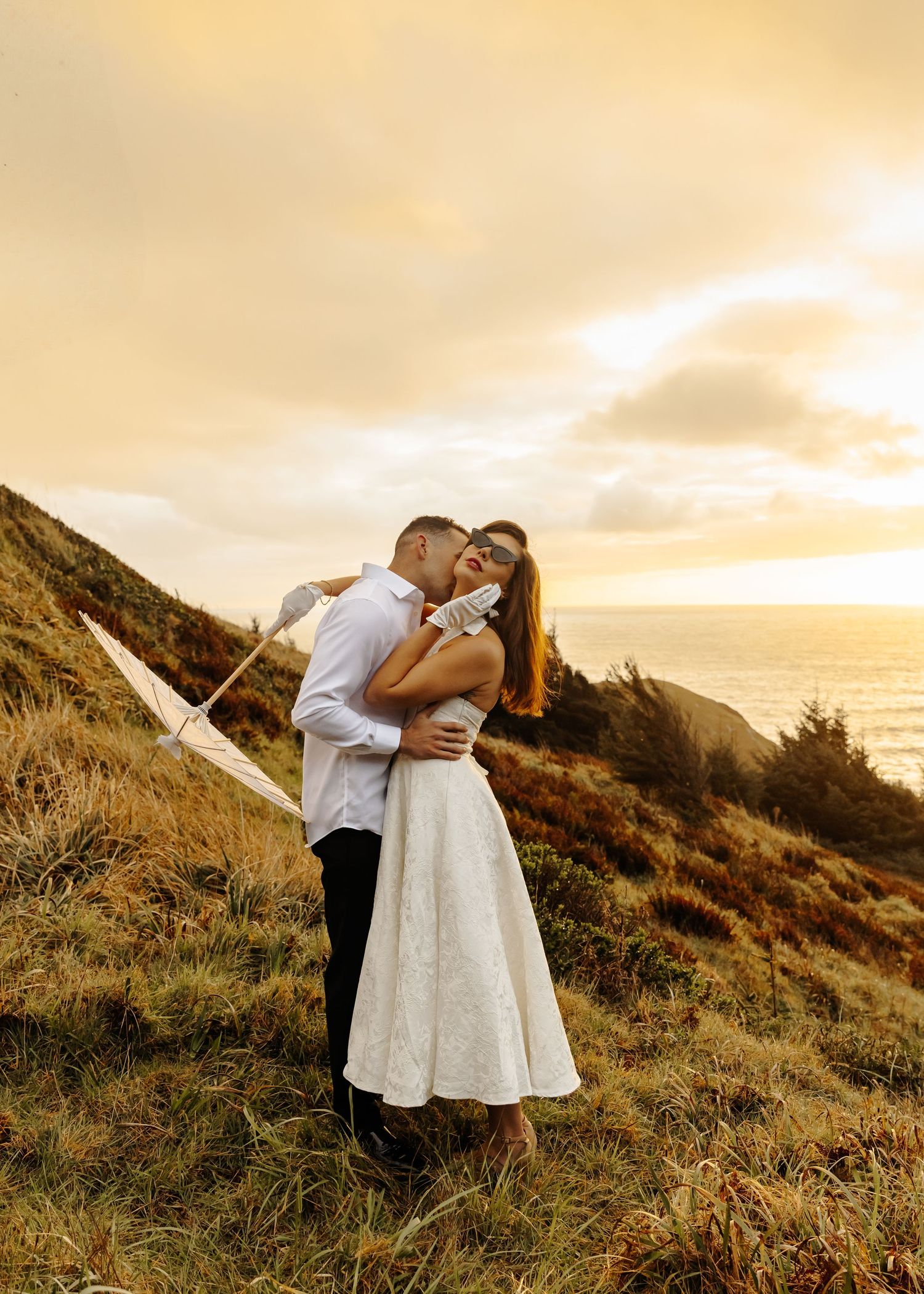 A romantic couple shares an intimate moment on a coastal hillside during golden hour sunset.