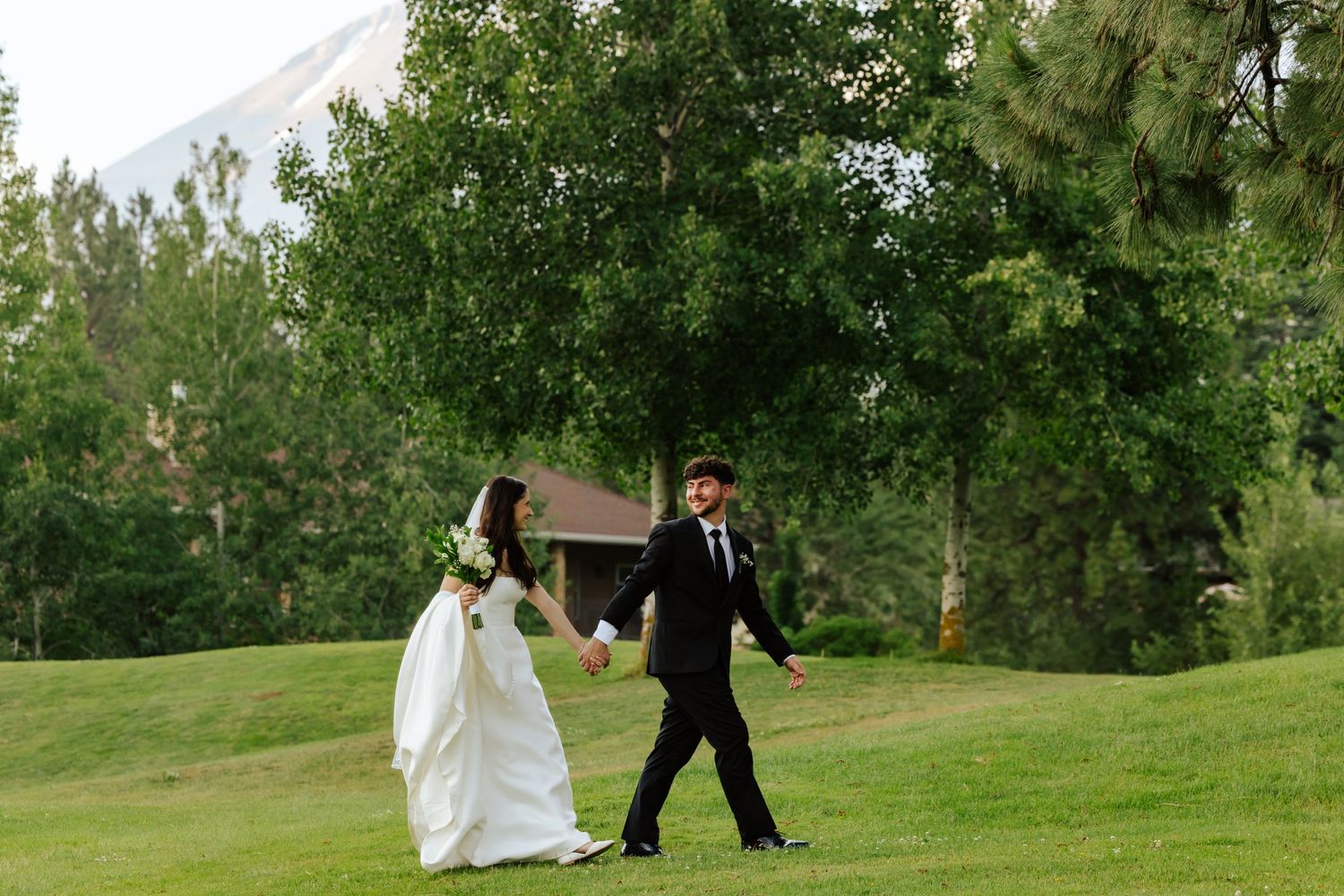Bride and groom walk hand in hand across lush green golf course with mountain and trees in background.