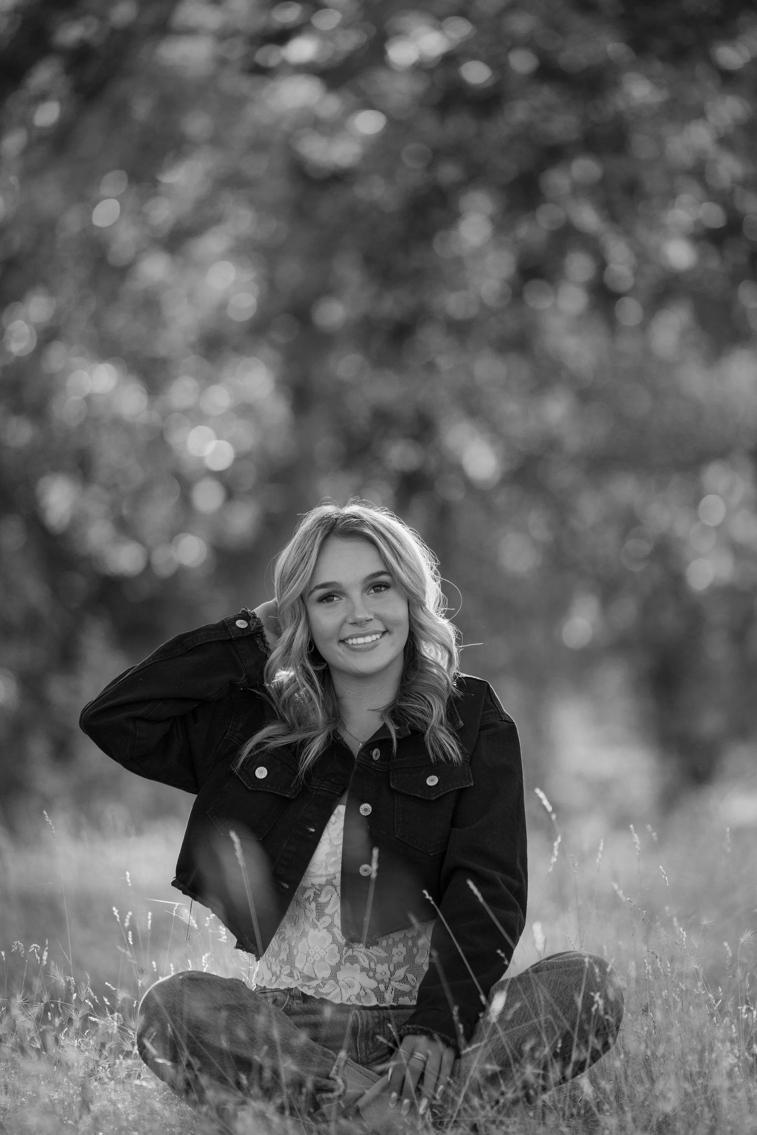 Person in denim jacket sits cross-legged in tall grass with bokeh background in monochrome.