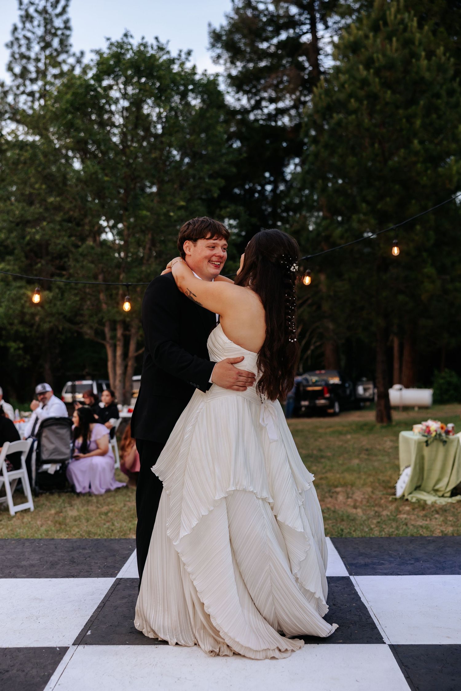 Bride and groom share first dance on checkered outdoor dance floor at dusk.