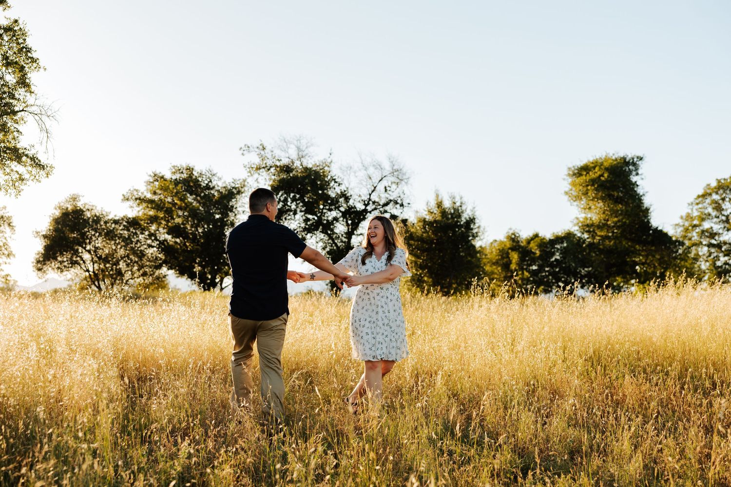 Couple holding hands and dancing in golden field with trees and clear blue sky during engagement photography session.
