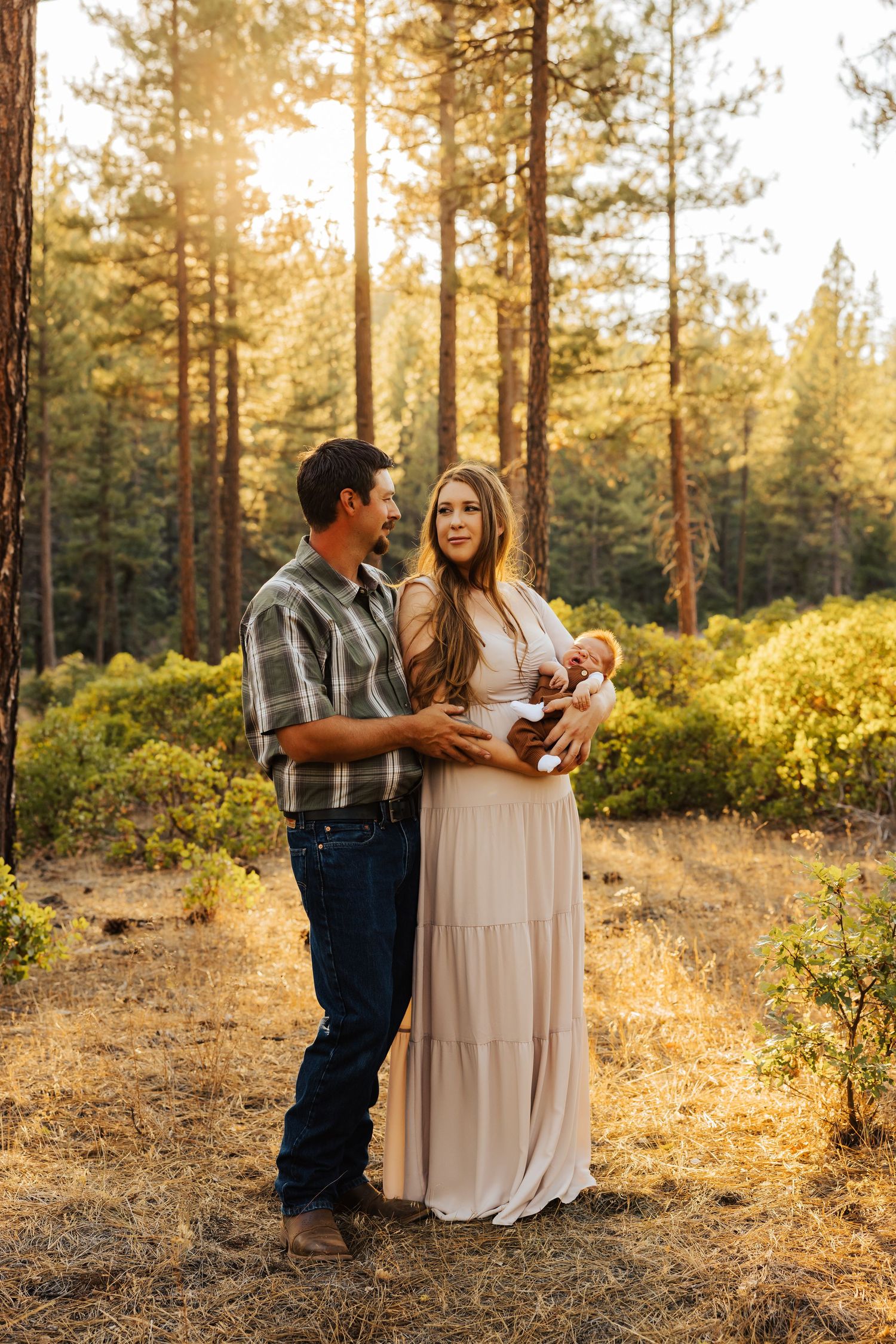 A couple holds their baby in a sunlit forest setting wearing casual western attire and a flowing dress.
