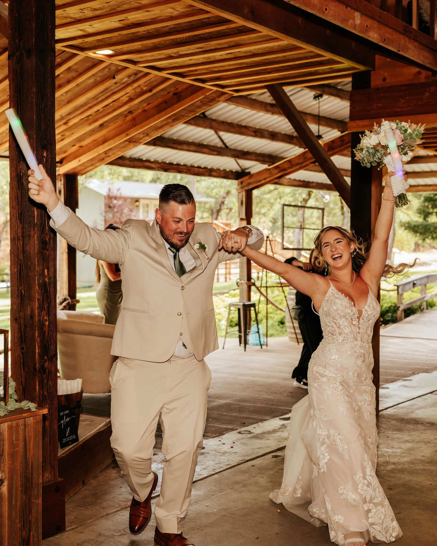 Wedding party celebrating with arms raised under wooden pavilion.