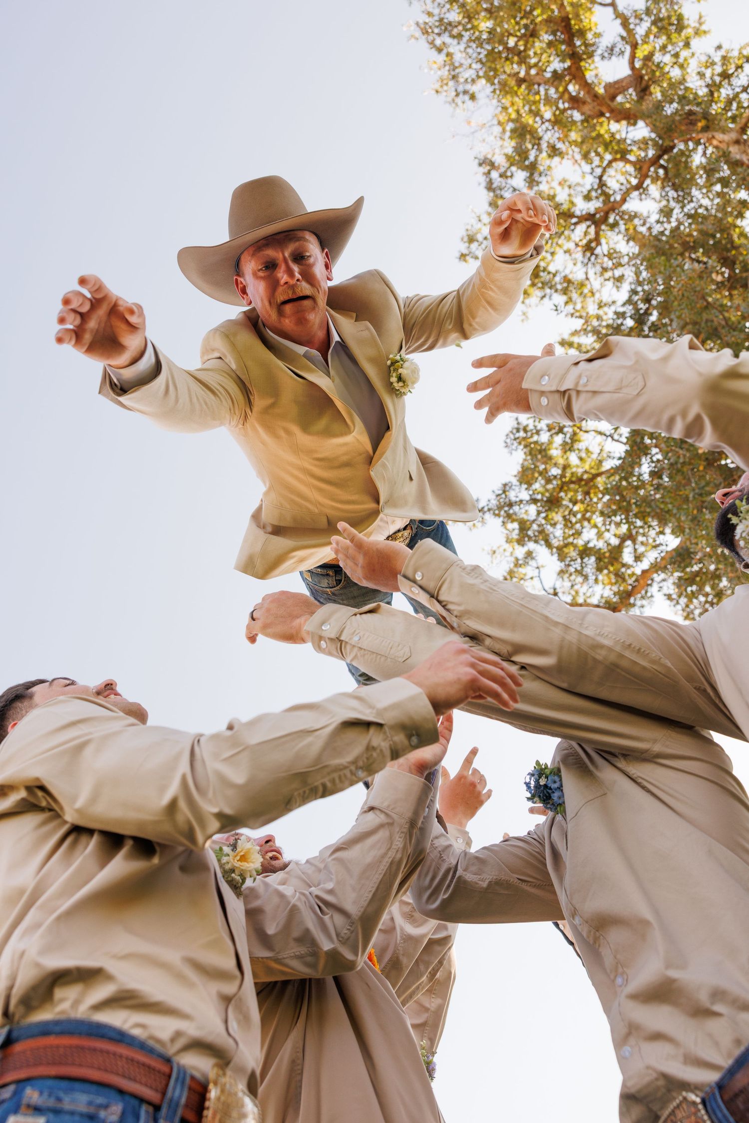 Groomsmen perform dynamic jumping poses in beige suits against blue sky.