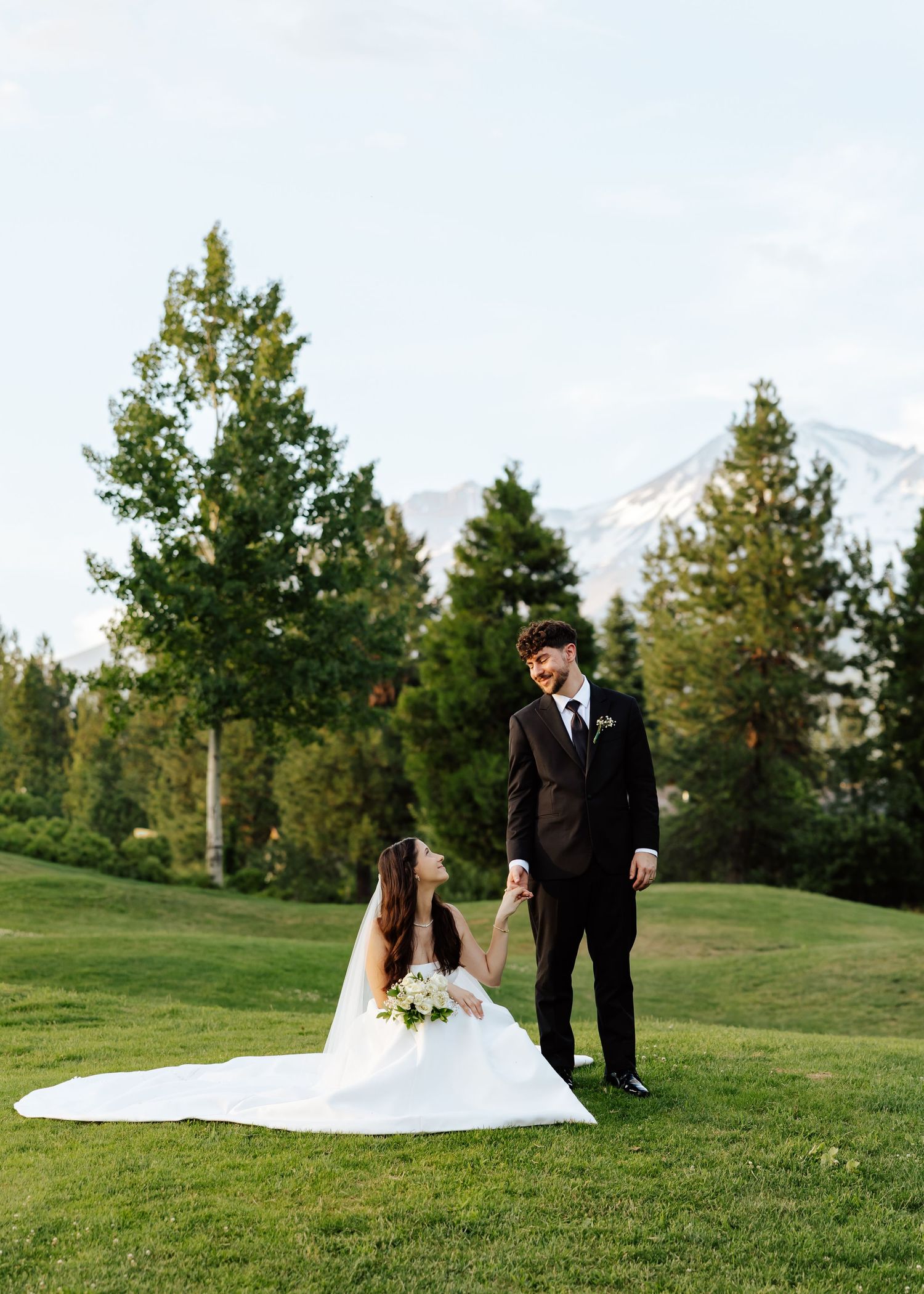 Bride in white gown and groom in black suit hold hands on green lawn with pine trees and snowy mountain backdrop.