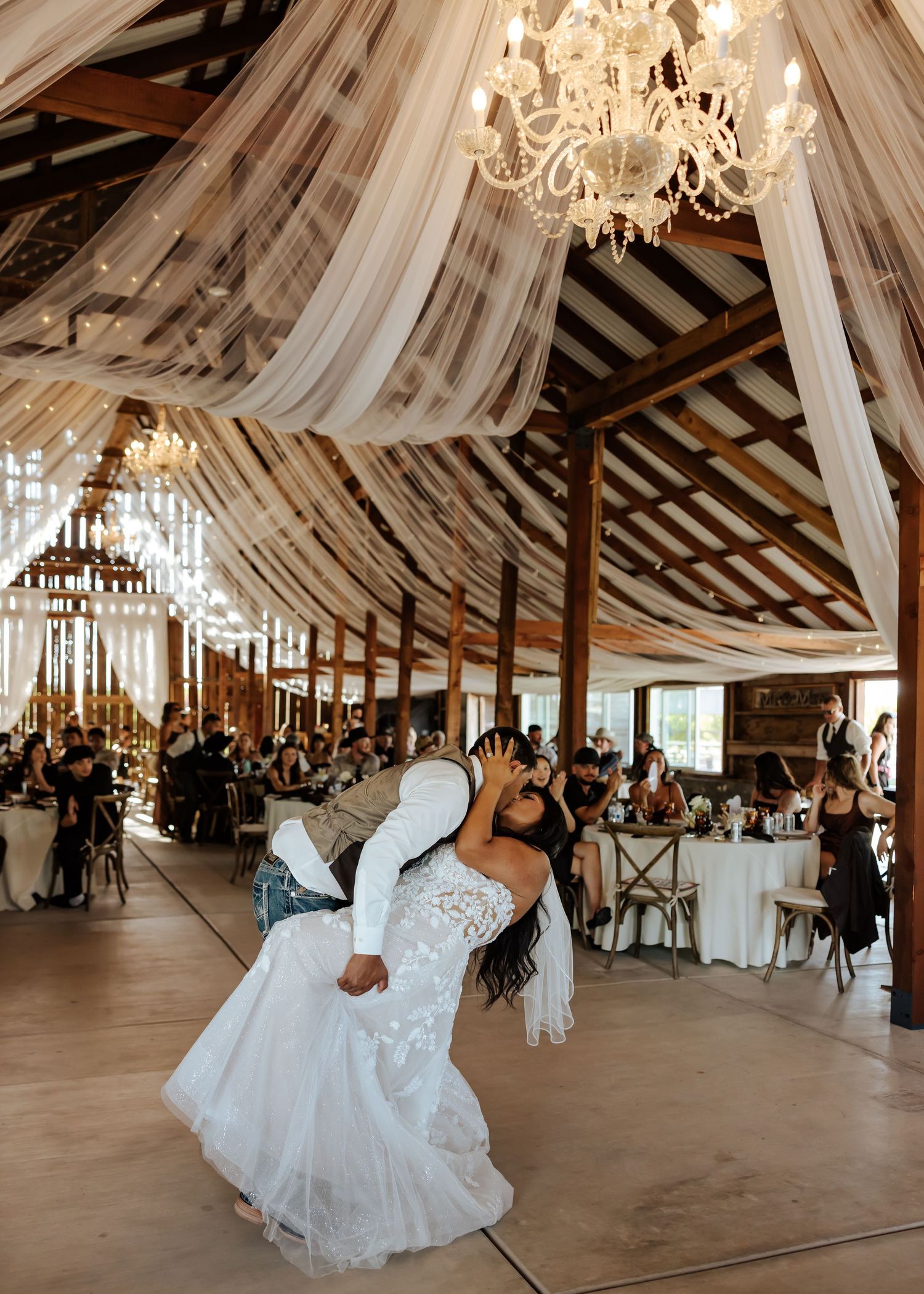 Bride and groom sharing first dance in a decorated barn venue with draped fabric ceiling and chandelier lighting.