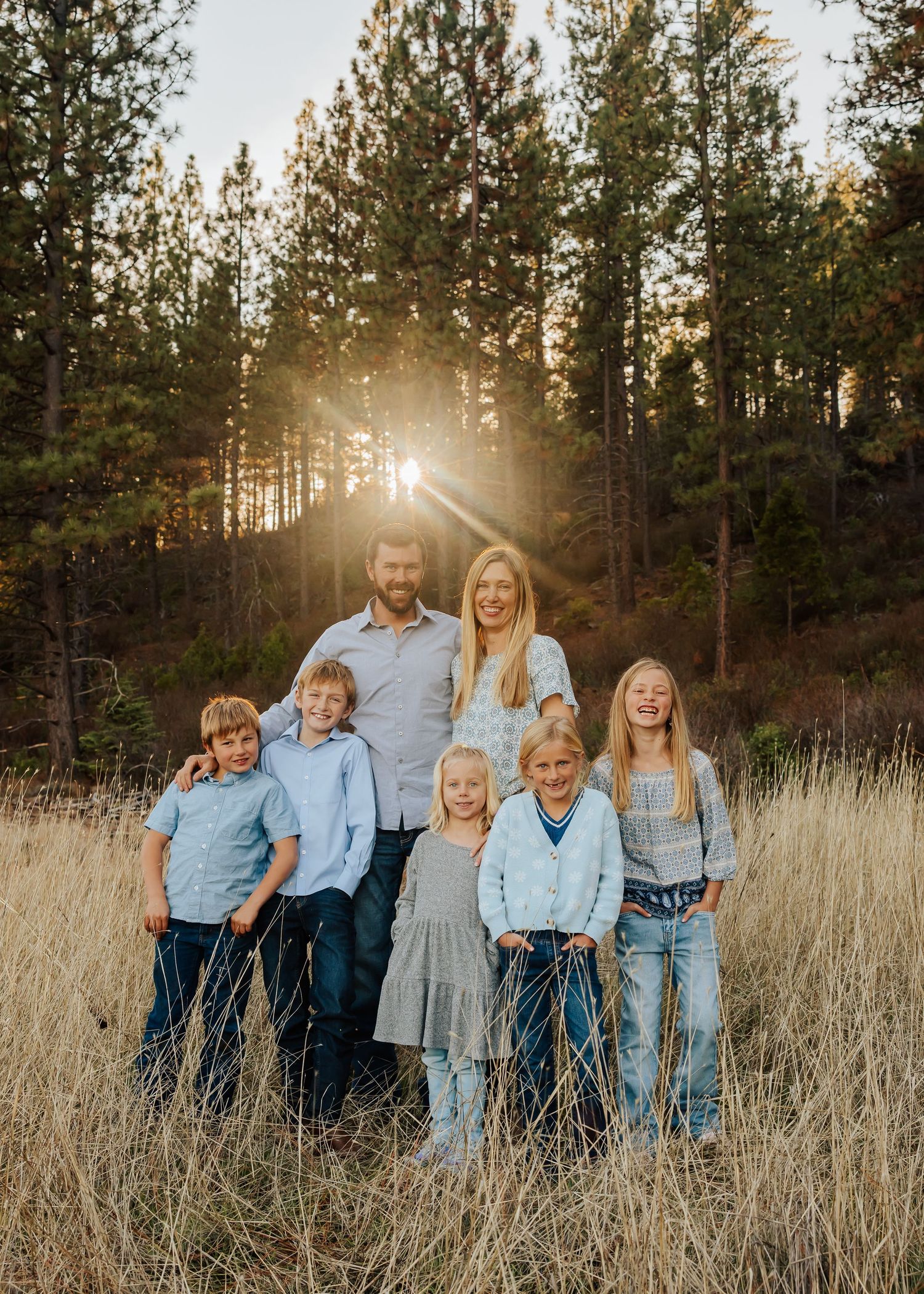 Extended family portrait in coordinating blue outfits posed in tall grass field at sunset.