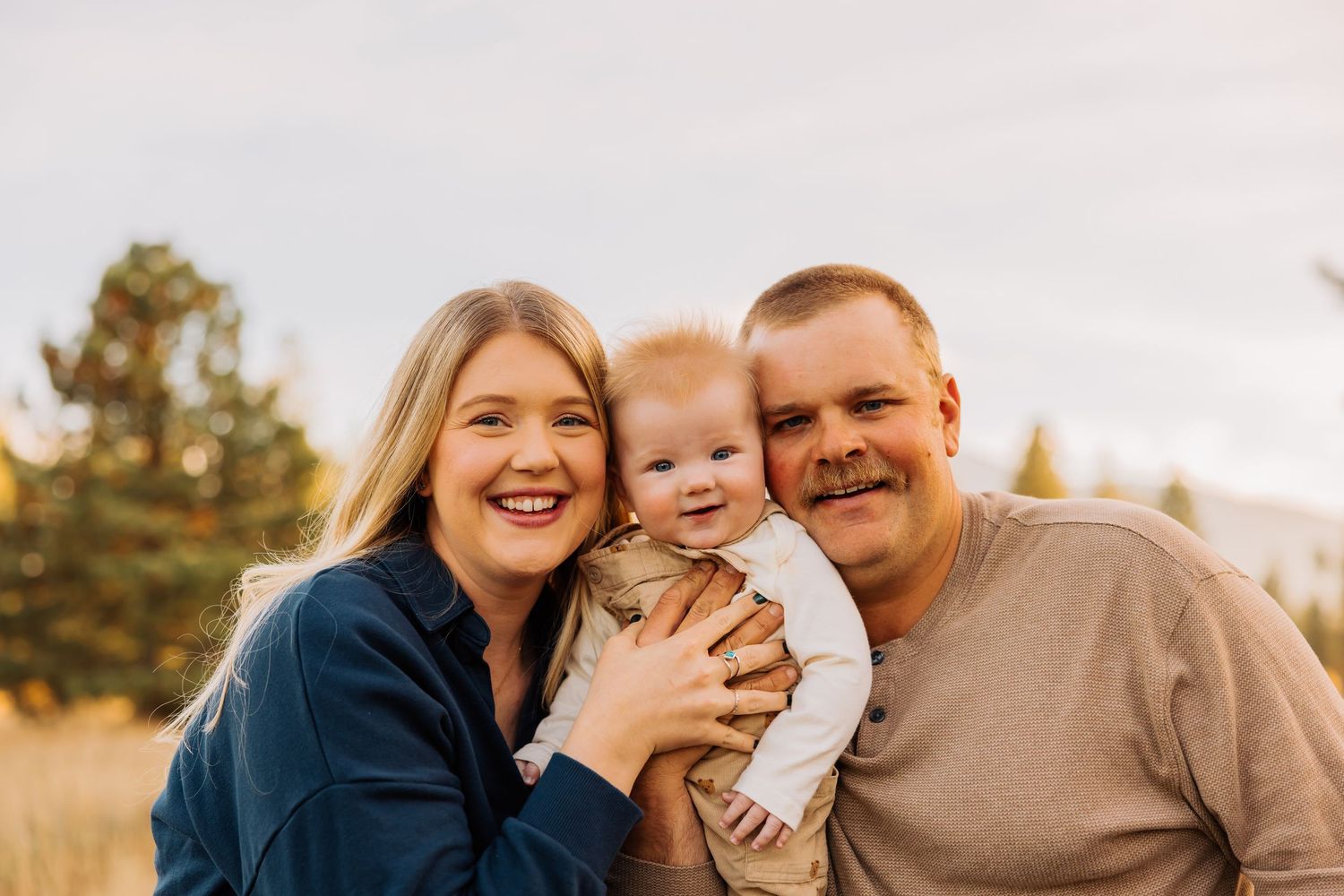 A happy family portrait in a golden field with evergreen trees and mountains in the distance.