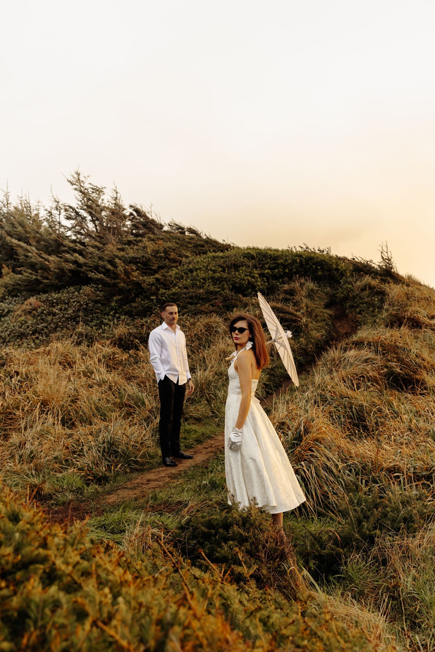 A couple in formal attire walks along a scenic coastal path with a white parasol.