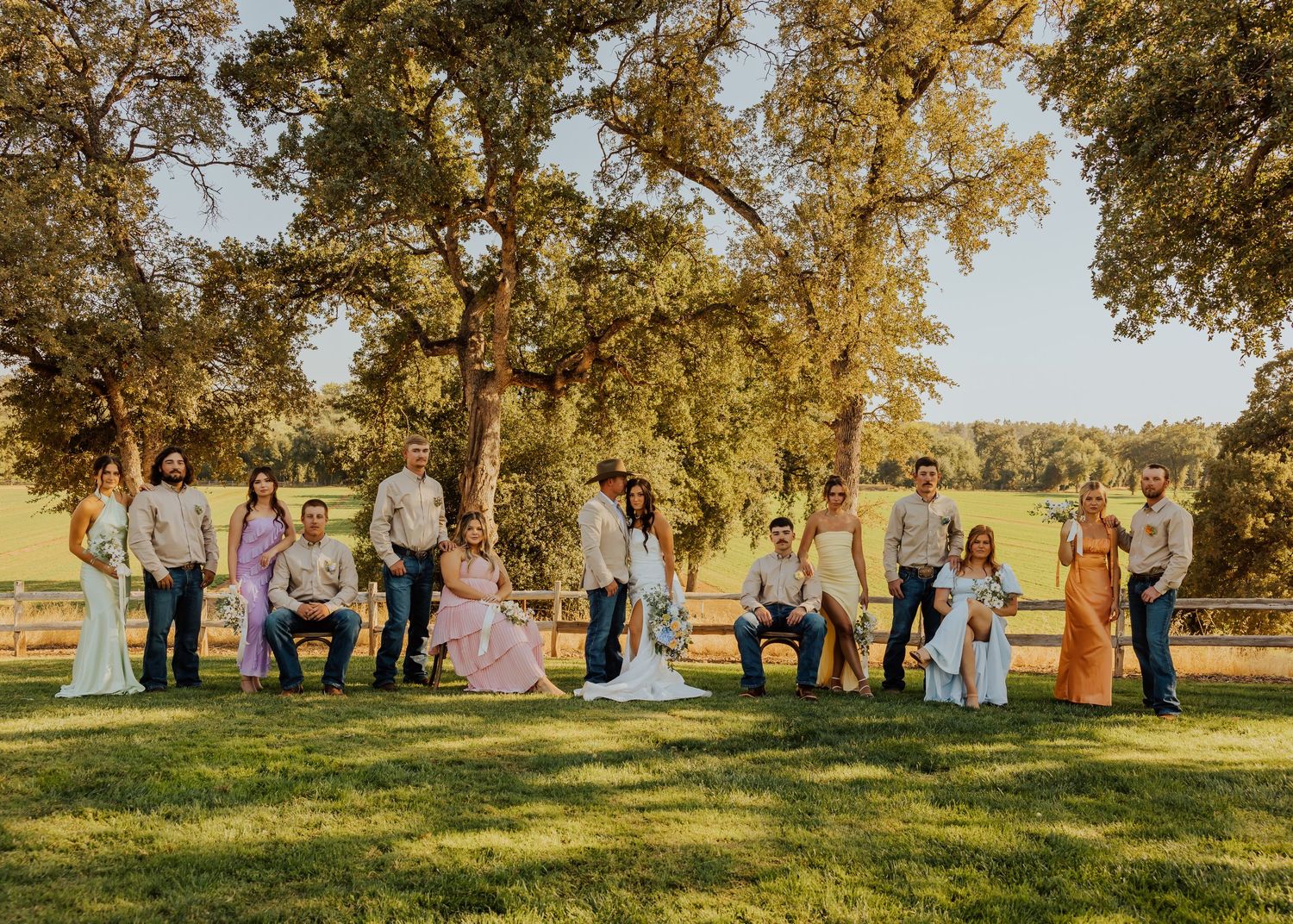 Wedding party in pastel attire poses under golden sunlit trees during sunset at an outdoor venue.