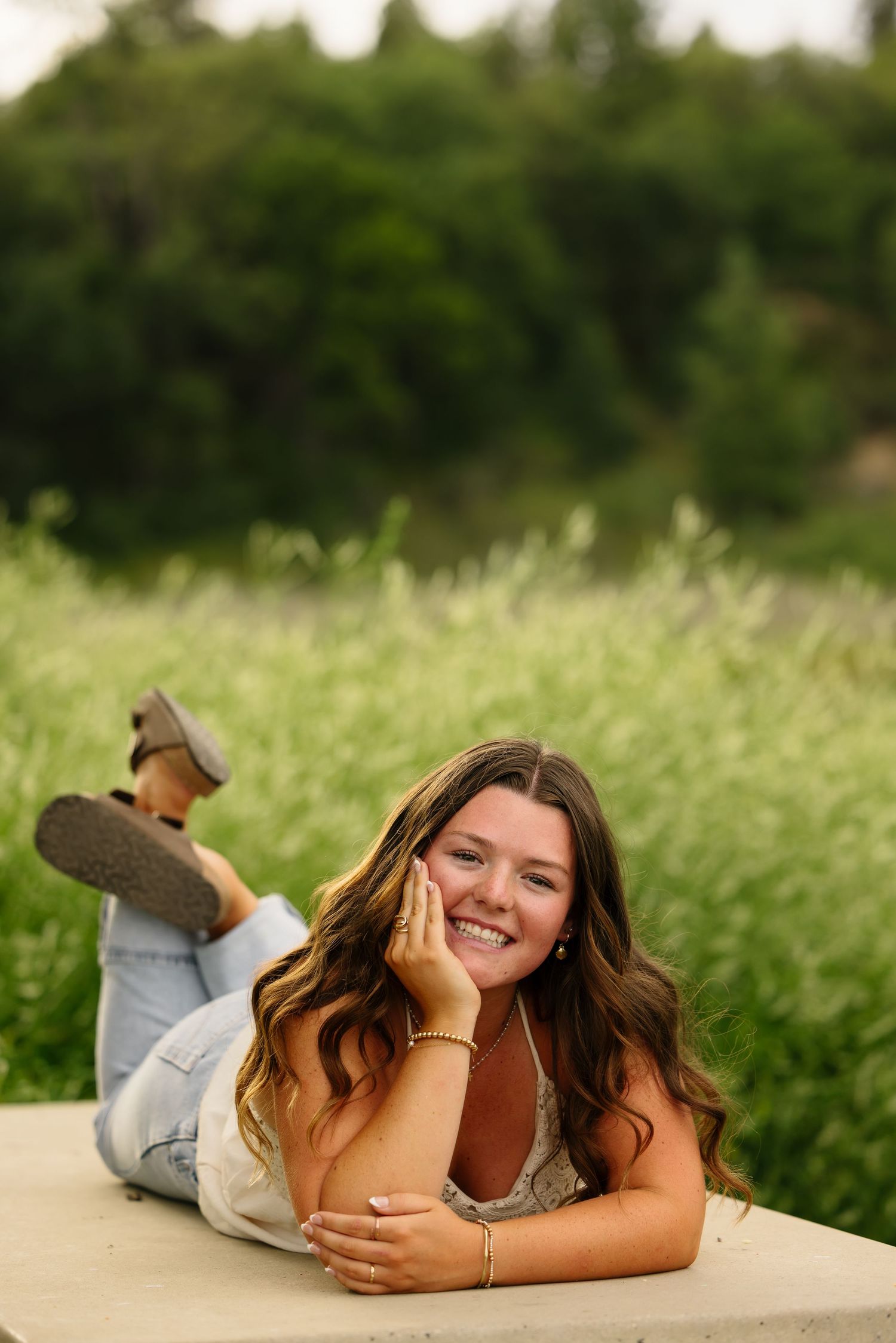 Young woman with long wavy hair lying on a ledge, smiling, wearing jeans in a lush green field.