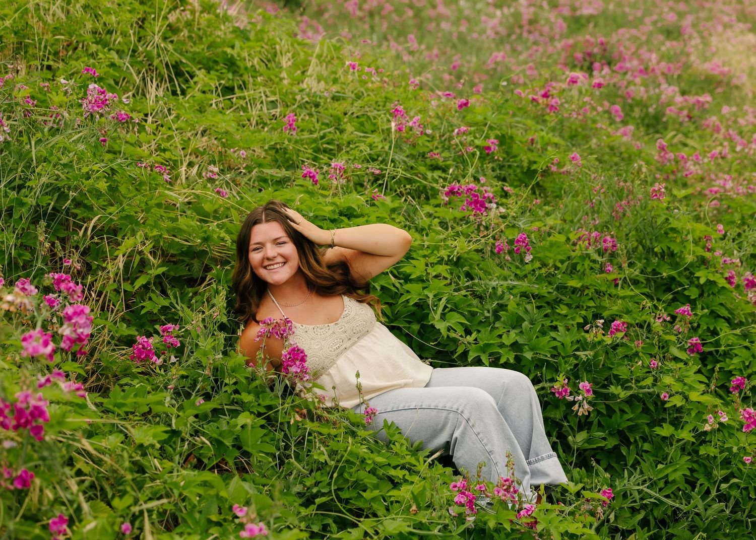Young woman smiling and relaxing in a lush green field of pink wildflowers on a bright summer day.