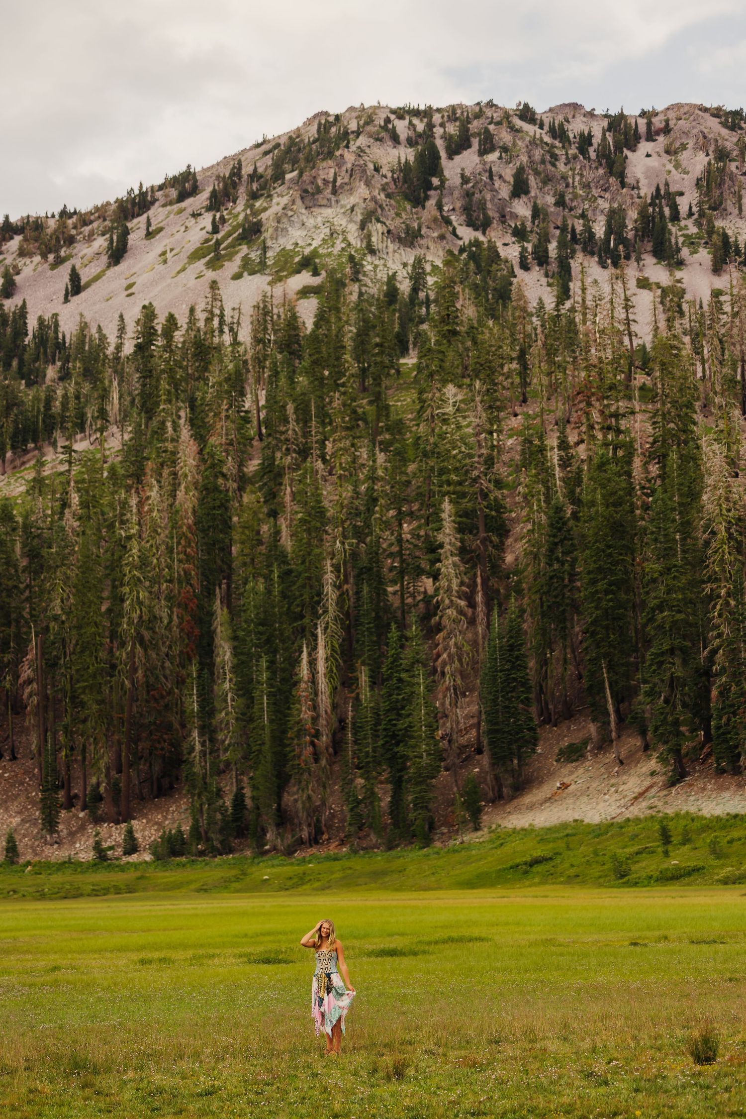 Woman in floral dress standing in green meadow with dense pine forest and rocky mountain peak in background.