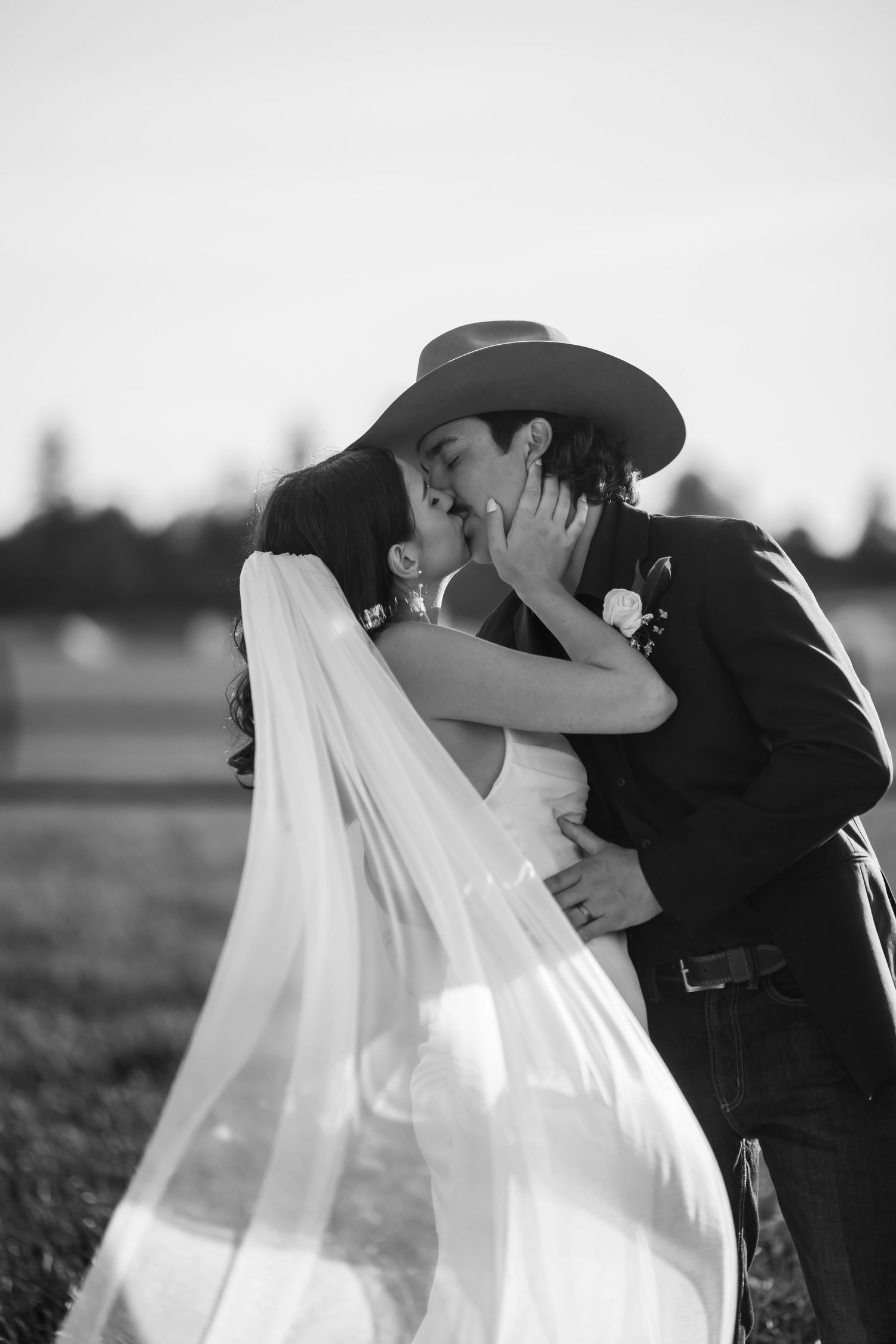 Romantic black and white wedding photo of couple kissing in a field with flowing veil.