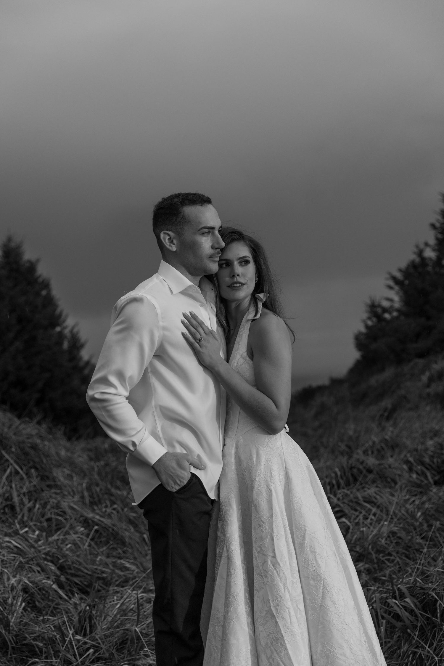 A romantic black and white portrait of a couple in formal wear on a hillside.