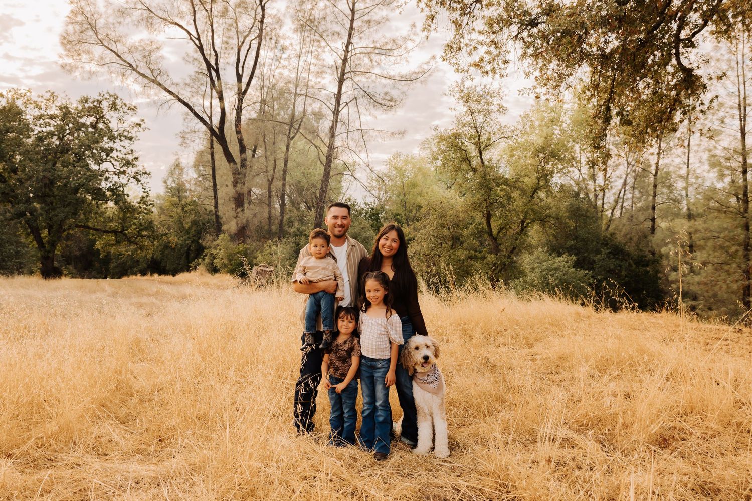 Group portrait in casual attire standing together in sunlit meadow with trees behind.