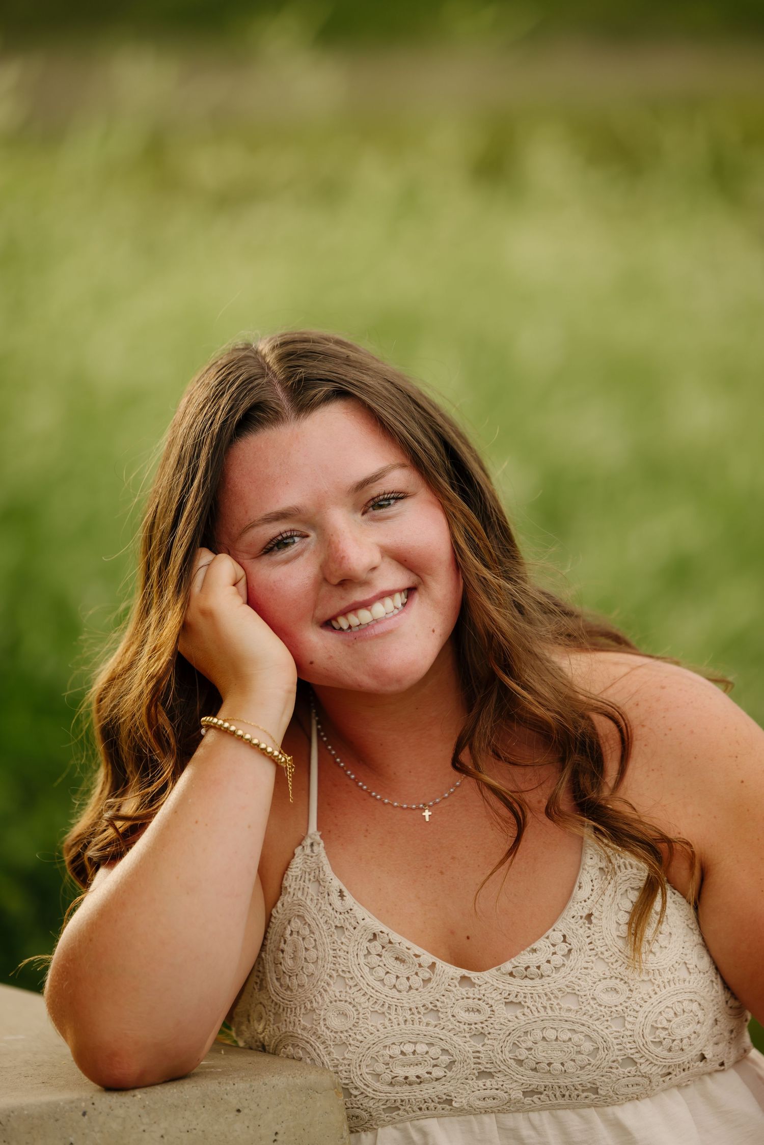Smiling young woman with wavy brown hair resting her cheek on her hand, wearing a lace top outdoors.