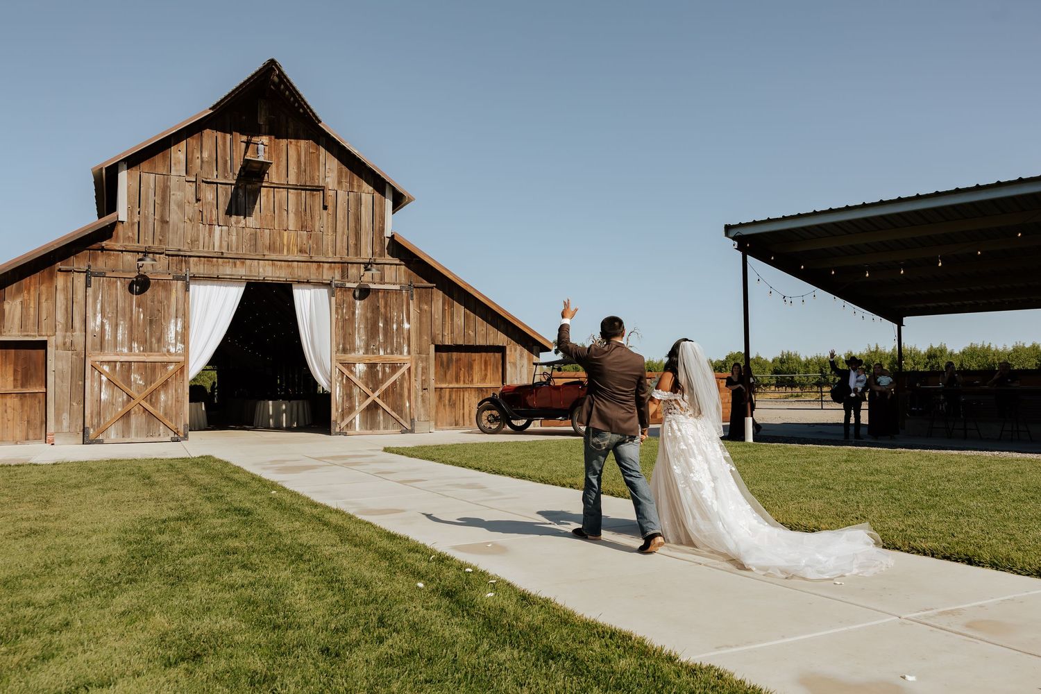 A bride and groom walk toward a rustic wooden barn wedding venue on a sunny day, with the bride's train trailing behind.