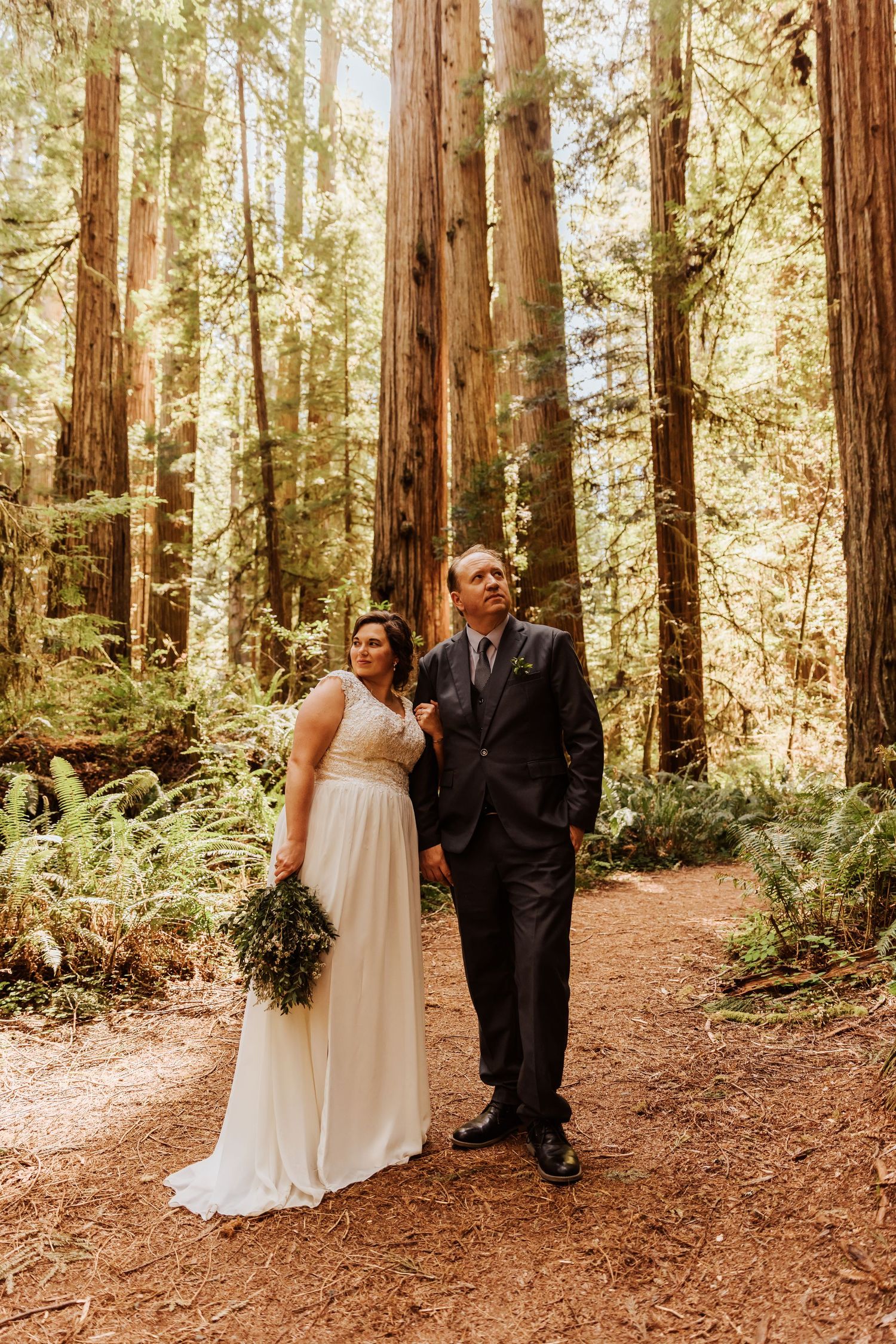 Couple celebrates their wedding day with playful moments in a sunlit redwood grove.