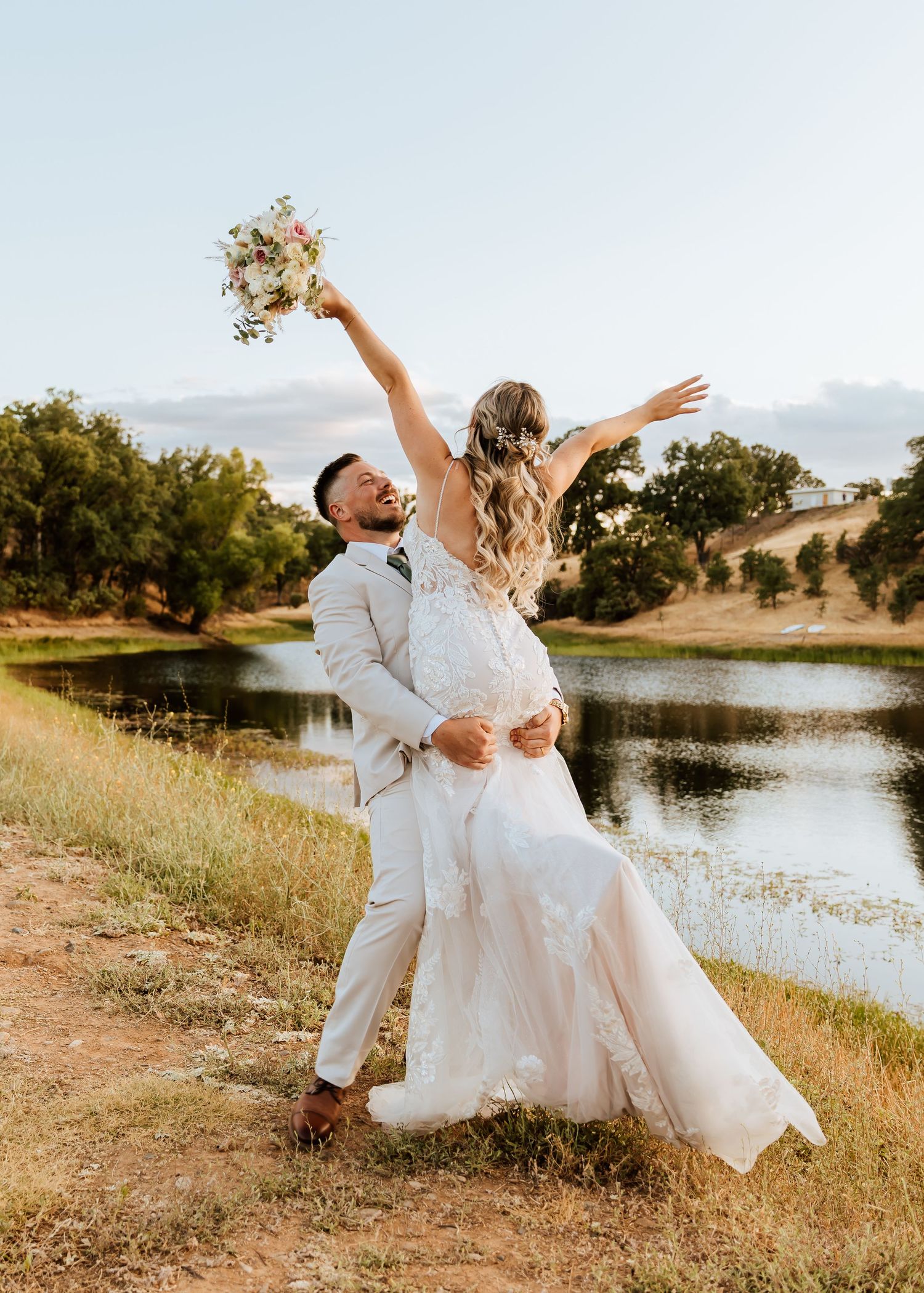 A joyful wedding couple celebrating with energetic poses by a lake during golden hour.