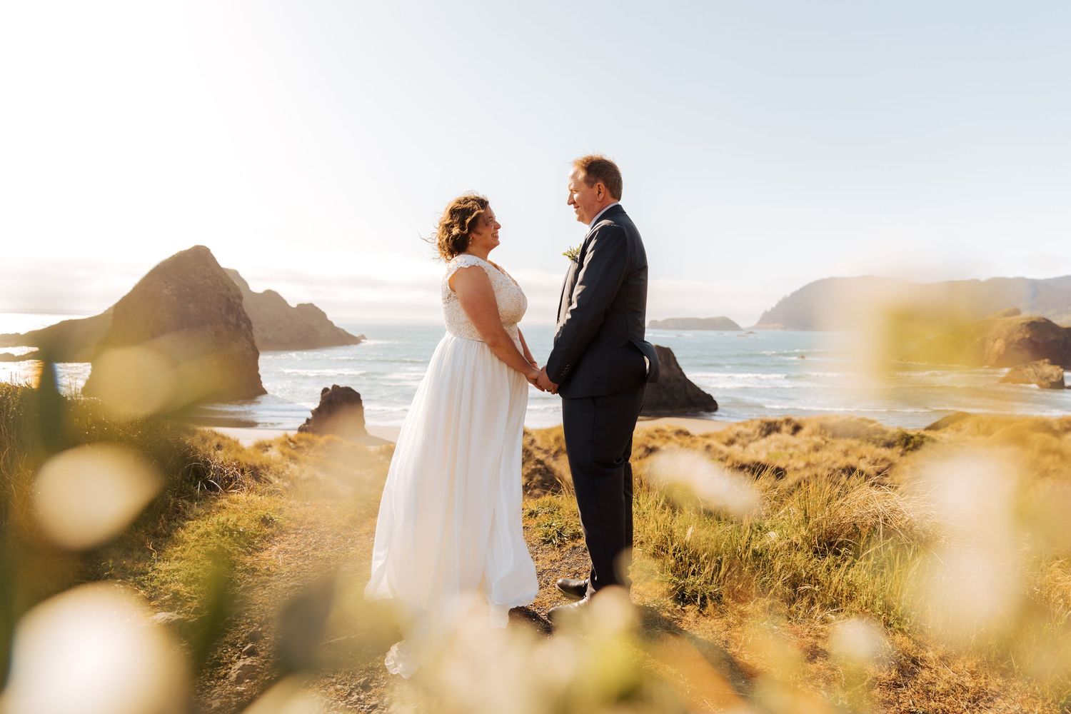 A couple embraces on a scenic coastal cliff during sunset with mountains and ocean in the background.