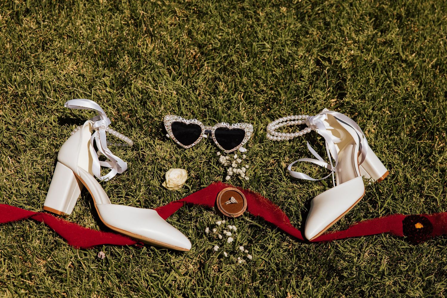 White wedding heels, heart-shaped sunglasses, and gold rings arranged on grass with red ribbon accents.