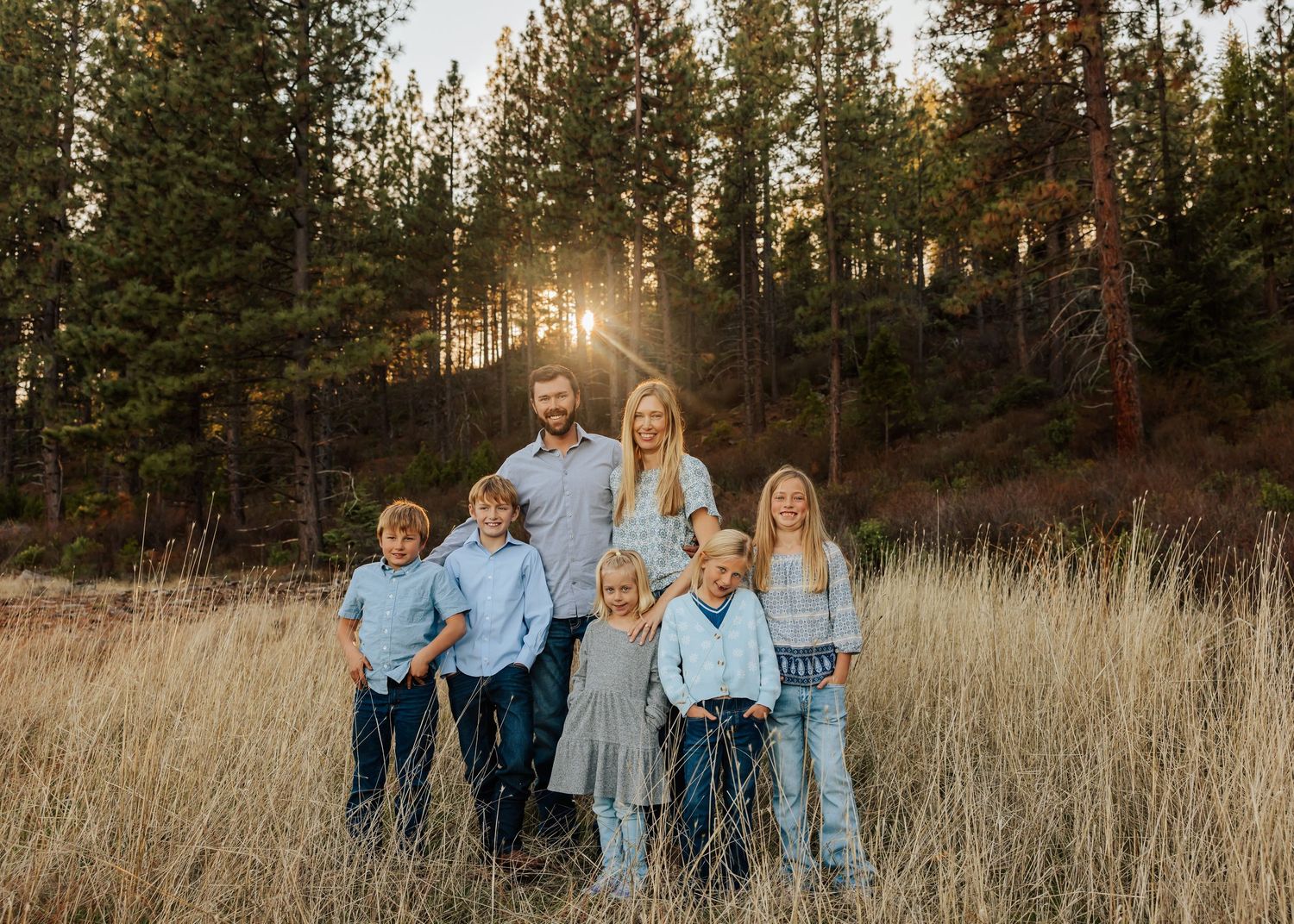 Family group photo session in casual denim and blue outfits against tall pine trees at sunset.