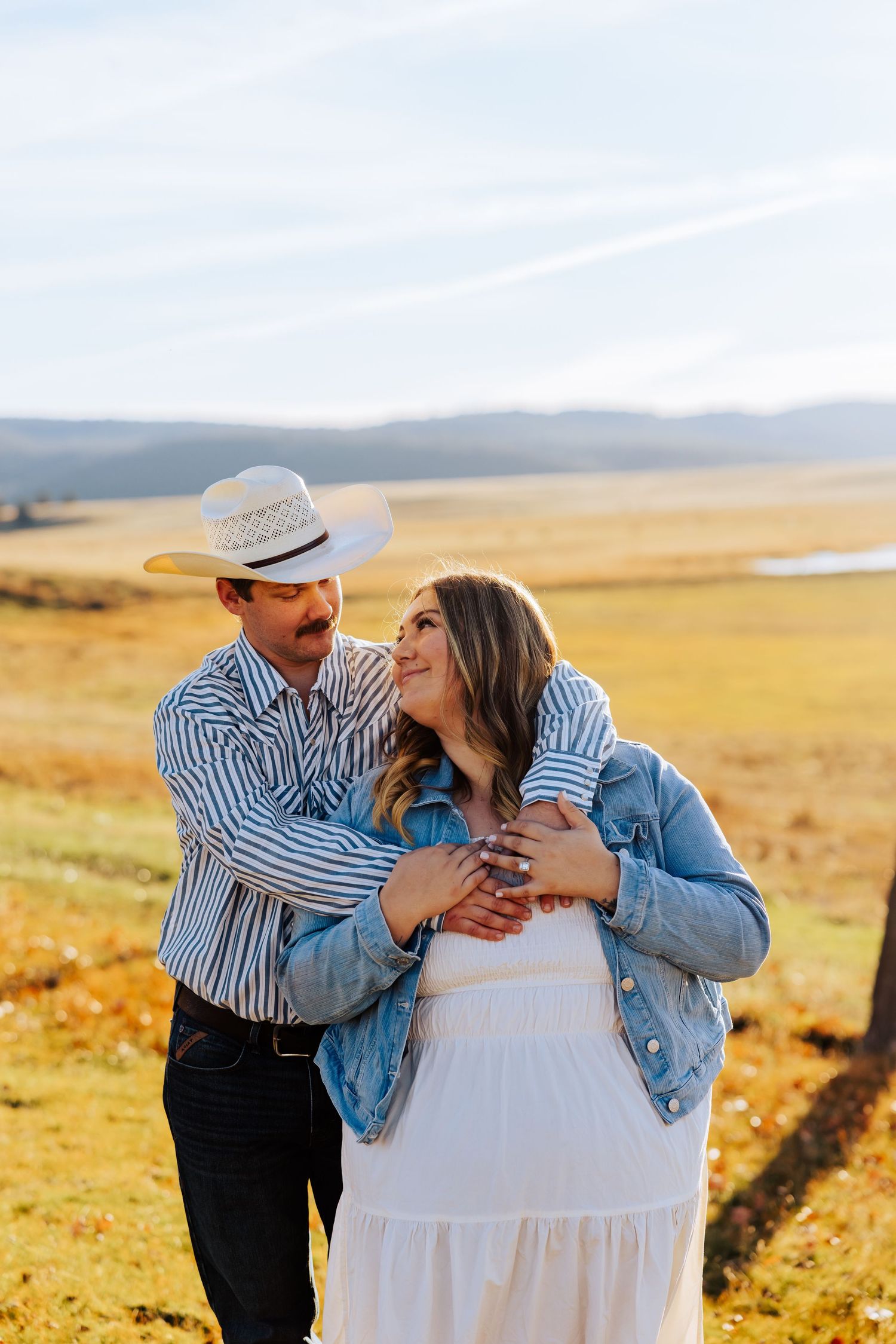 A couple shares loving embrace in sunlit field with mountains in background.